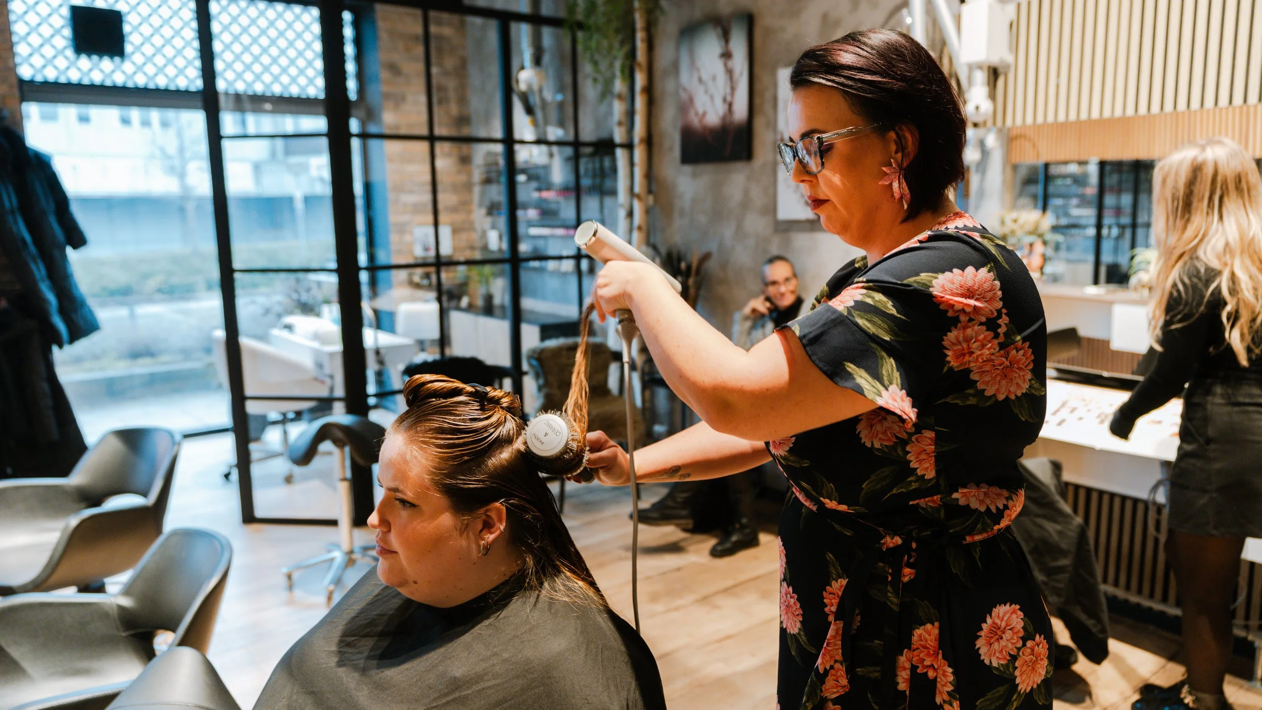 Kvinde får føntørring i frisørsalon, med flere personer i baggrunden