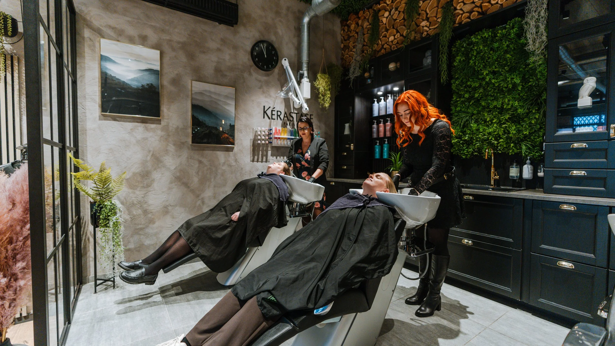 To kvinder får vasket håret i en salon, mens to personer ser til. En medarbejder med rødt hår skyller deres hår, og en anden medarbejder står bag dem.