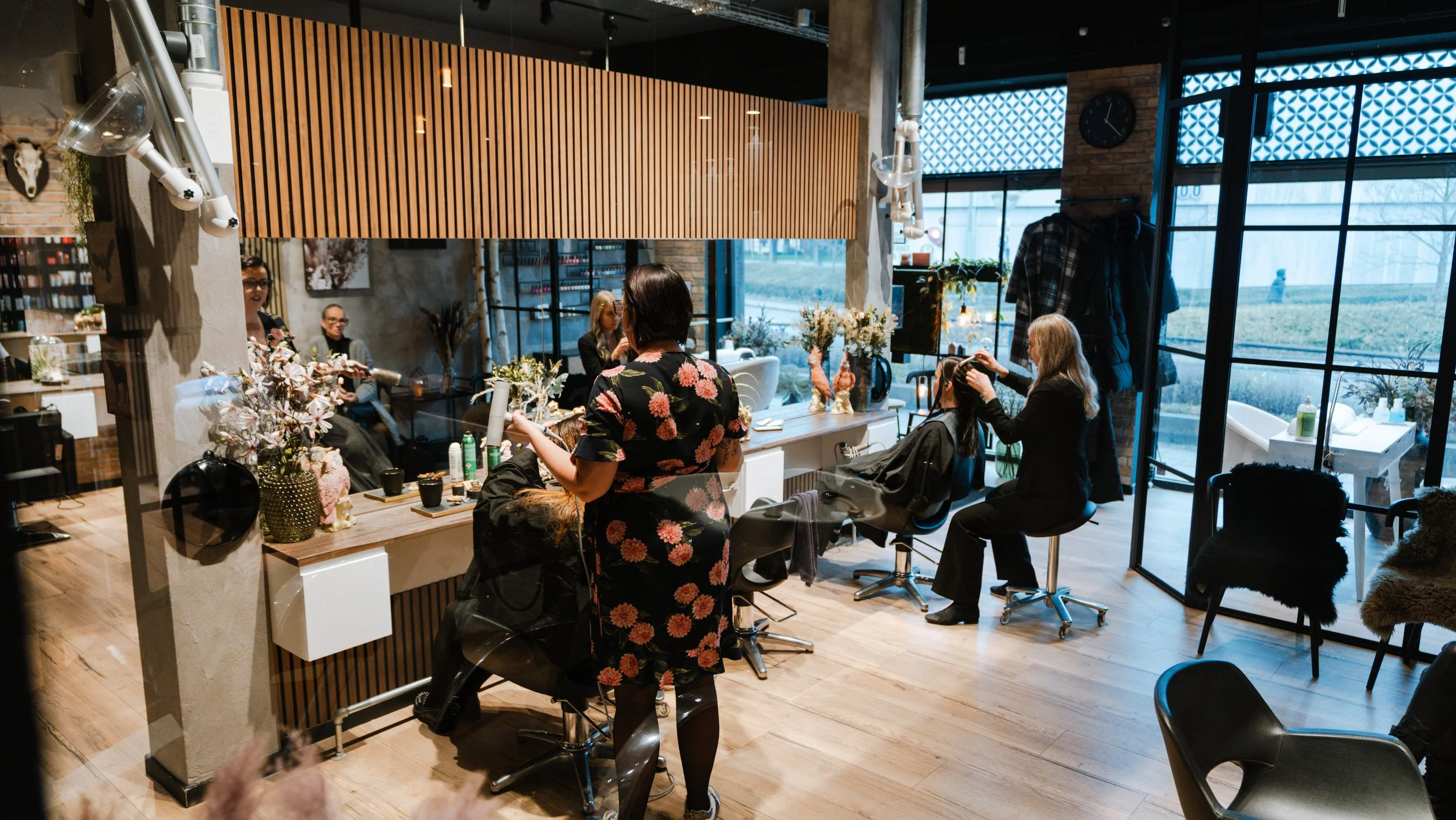 Kvindelig frisør klipper en kundes hår i en moderne frisørsalon med store vinduer og trædetaljer.