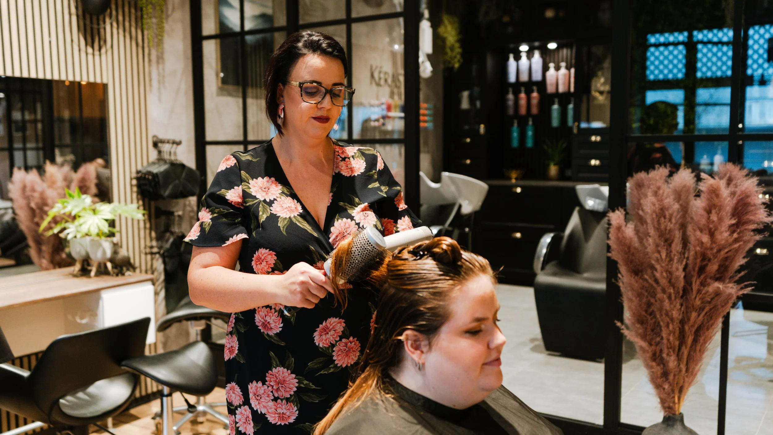 En frisør, en kvinde med kort mørkt hår og store briller, vasker et andet kvindes hår med føntørrer i en moderne frisørsalon.