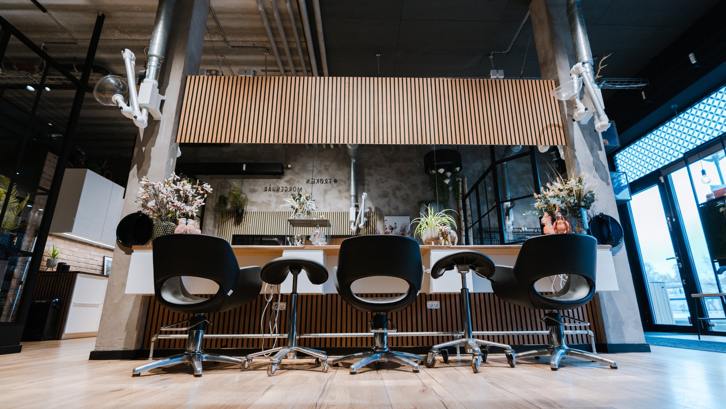 Indendørs kontor med tre sorte kontorstole foran en lang hvid bordplade, dekoreret med blomster og planter. Bagved er der en opadgående trævæg og store glasdøre til udendørs område.