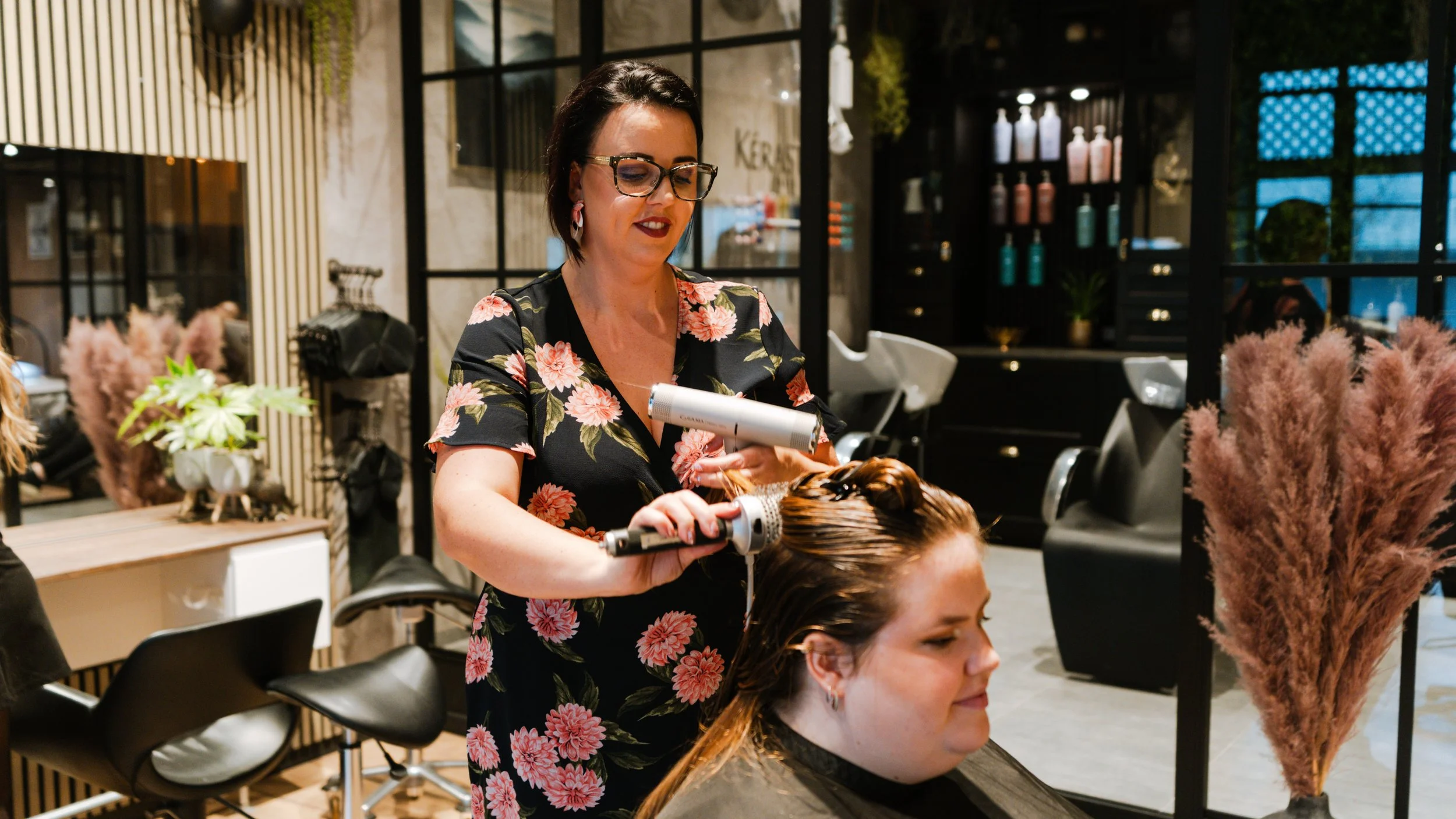 En frisør, der bruger føntørre til at style en kvindes hår, mens de sidder i en moderne frisørsalon.