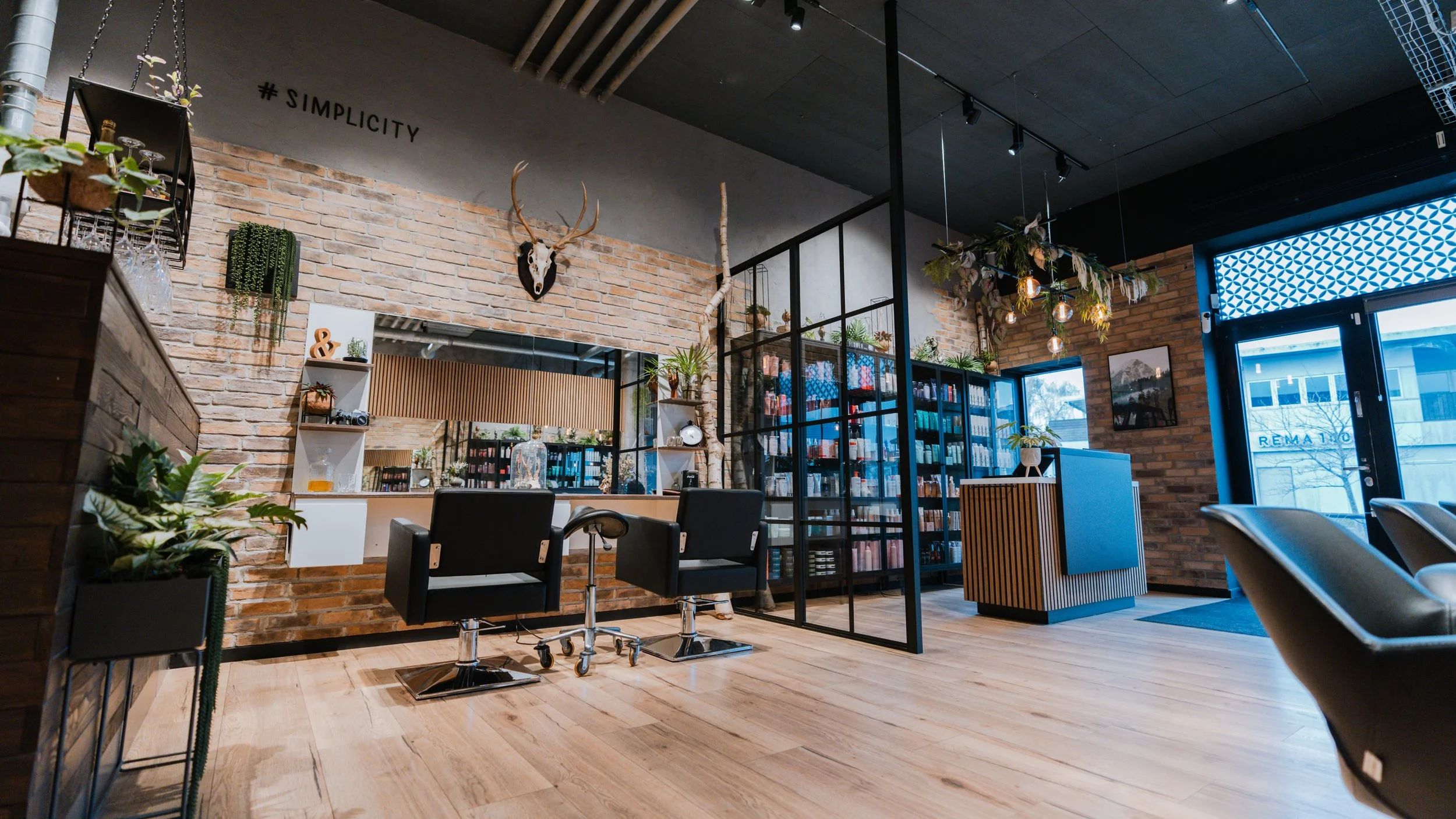 Indretning af et moderne frisørsalon med læderstole, plantepynt, brændt murstensvæg med decorative elementer som et hjortehoved, og store vinduer. Der er hylder med skjønt, og en check-in disk.