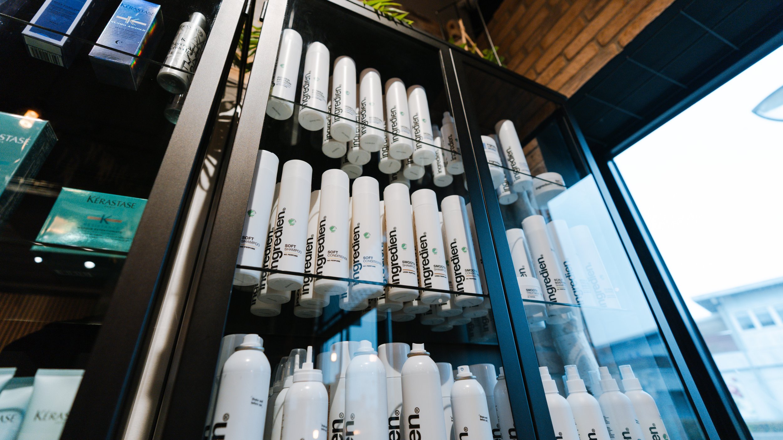 Reol med hvide hårplejeprodukter fra mærket Ingredien, plassert i en butik, set igennem en glaslåge . Der er også nogle blå Kérastase produkter i baggrunden.