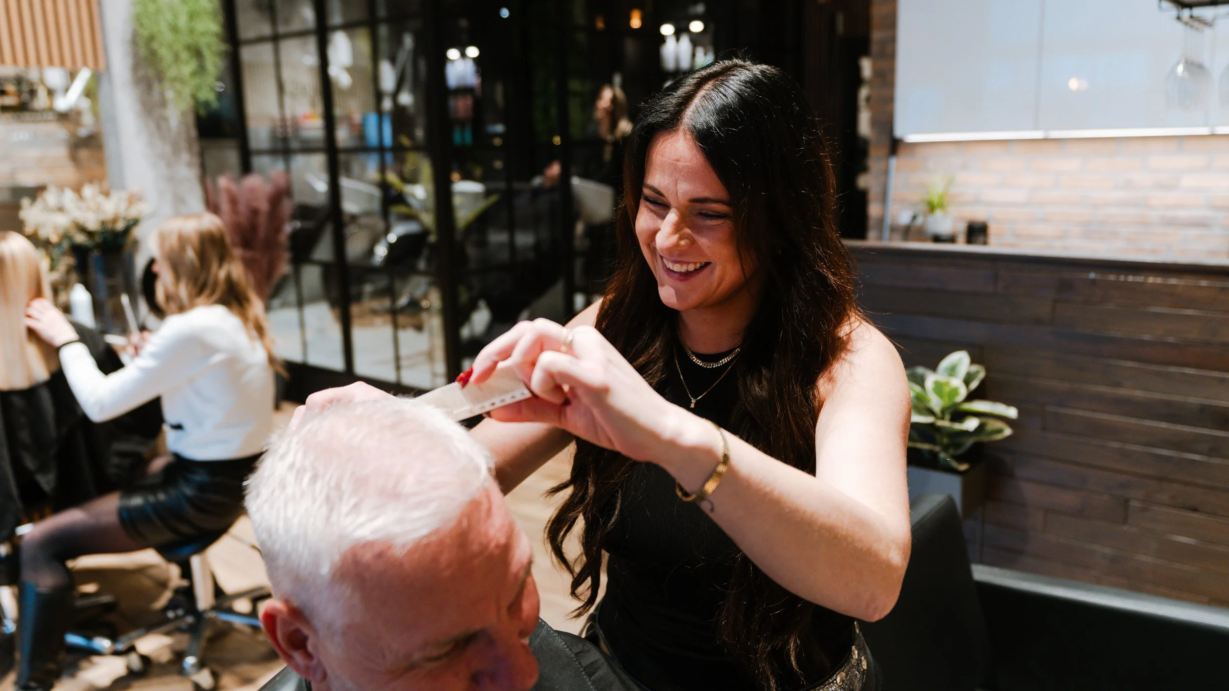 Kvinde med langt mørkt hår smiler, mens hun klipper en mand i en frisørsalon.