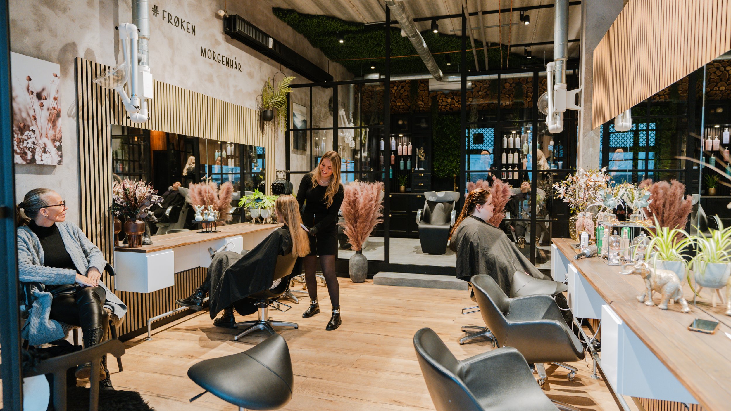 Indretning af en frisør salon med kunder og frisører, grønne planter, dekorative blomster, og store spejle.