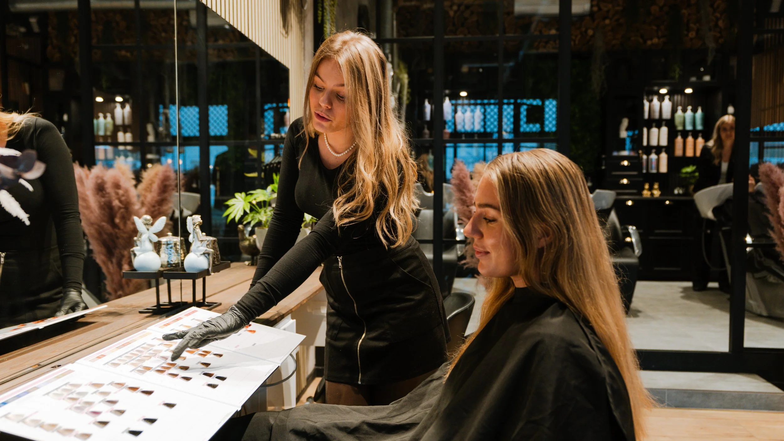 To kvinder i en frisørsalon, der vælger hårfarver fra farvekort, med spejl og greenery i baggrunden.