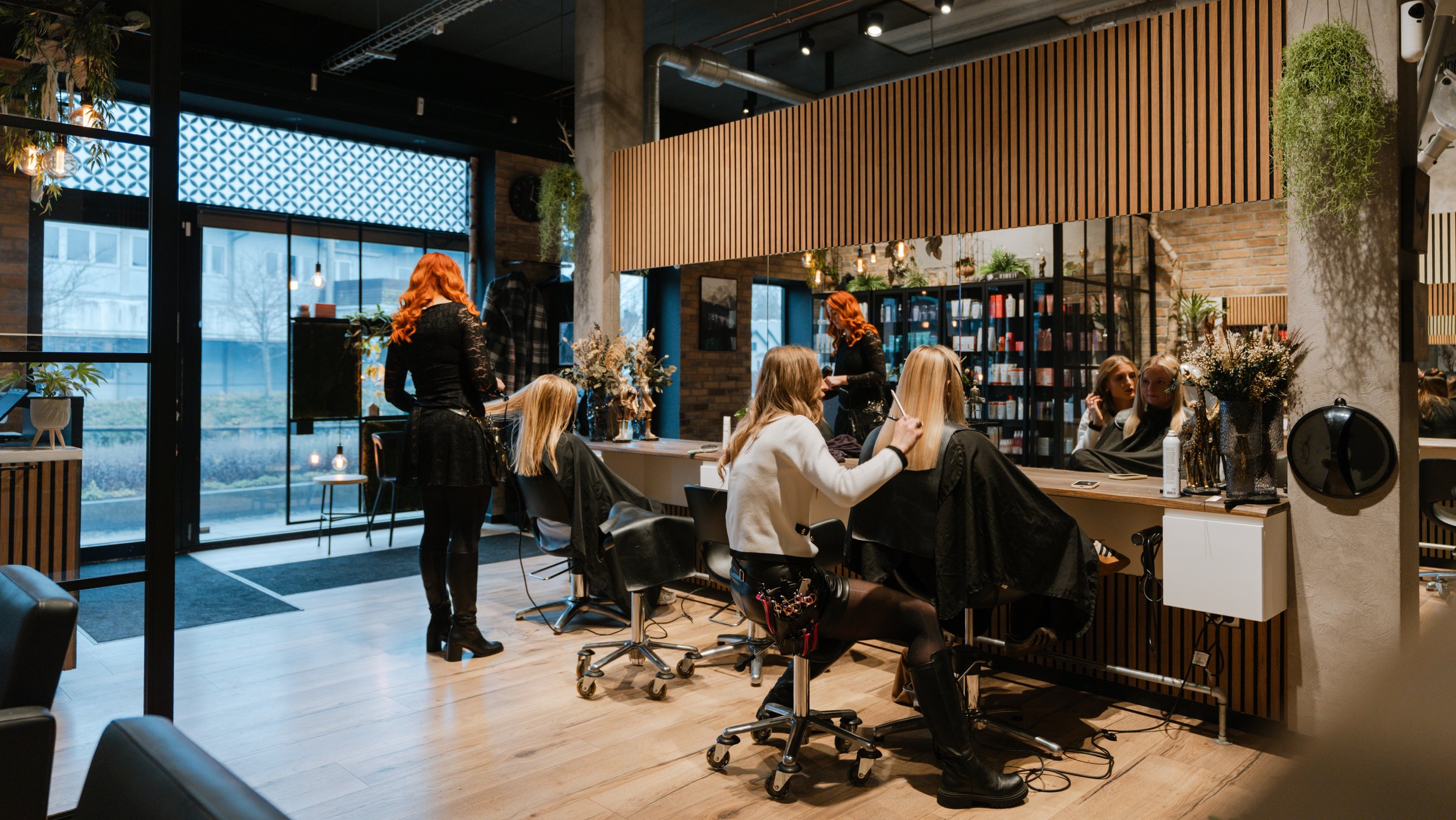En frisørsalon med flere kvinder, der får klippet hår, og en frisør med rødt hår, som arbejder bagved.