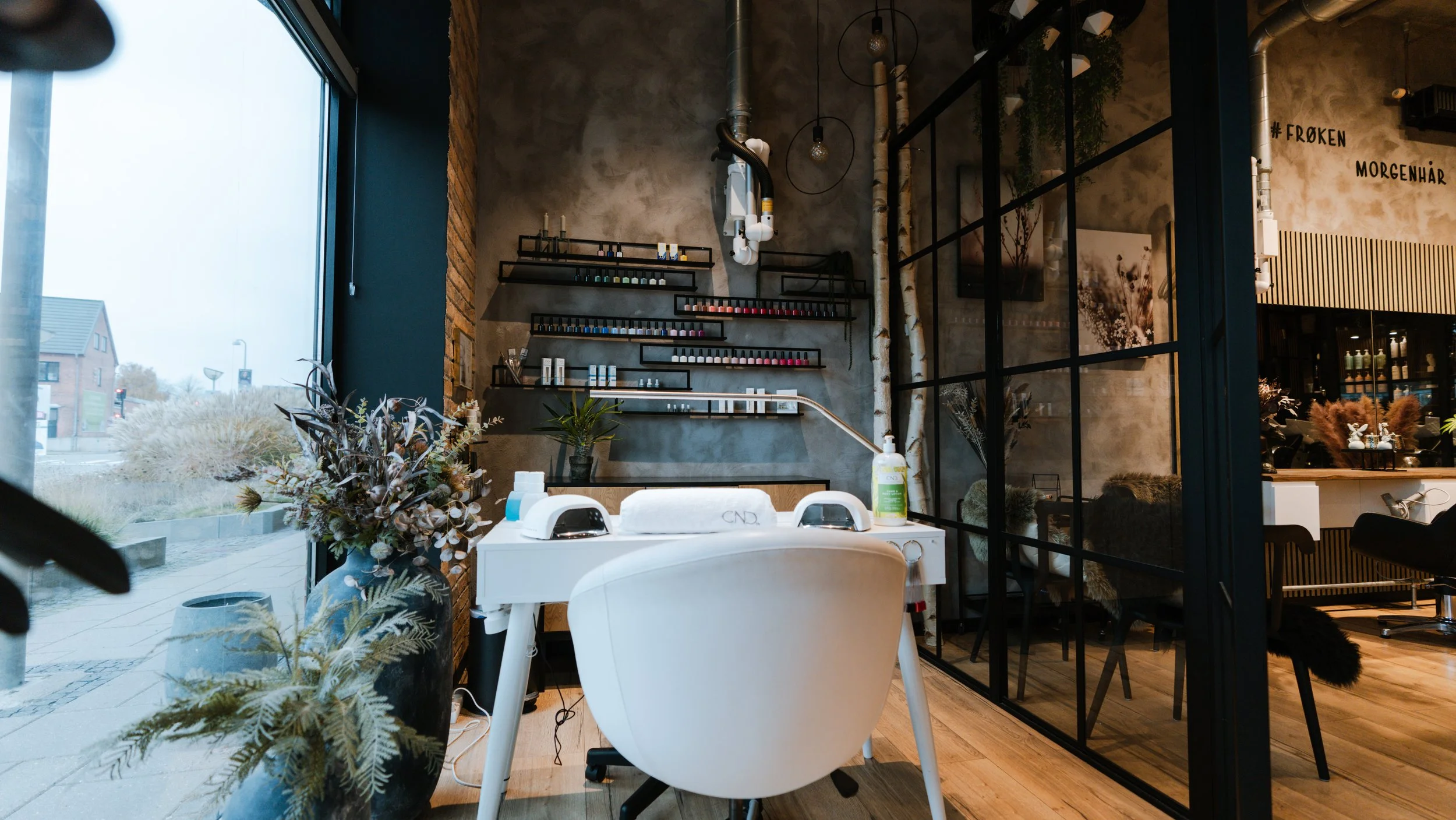 Indendørs frisørsalon med manicure-arbejdsstation, botaniske dekorationer og væg med behandlingsprodukter