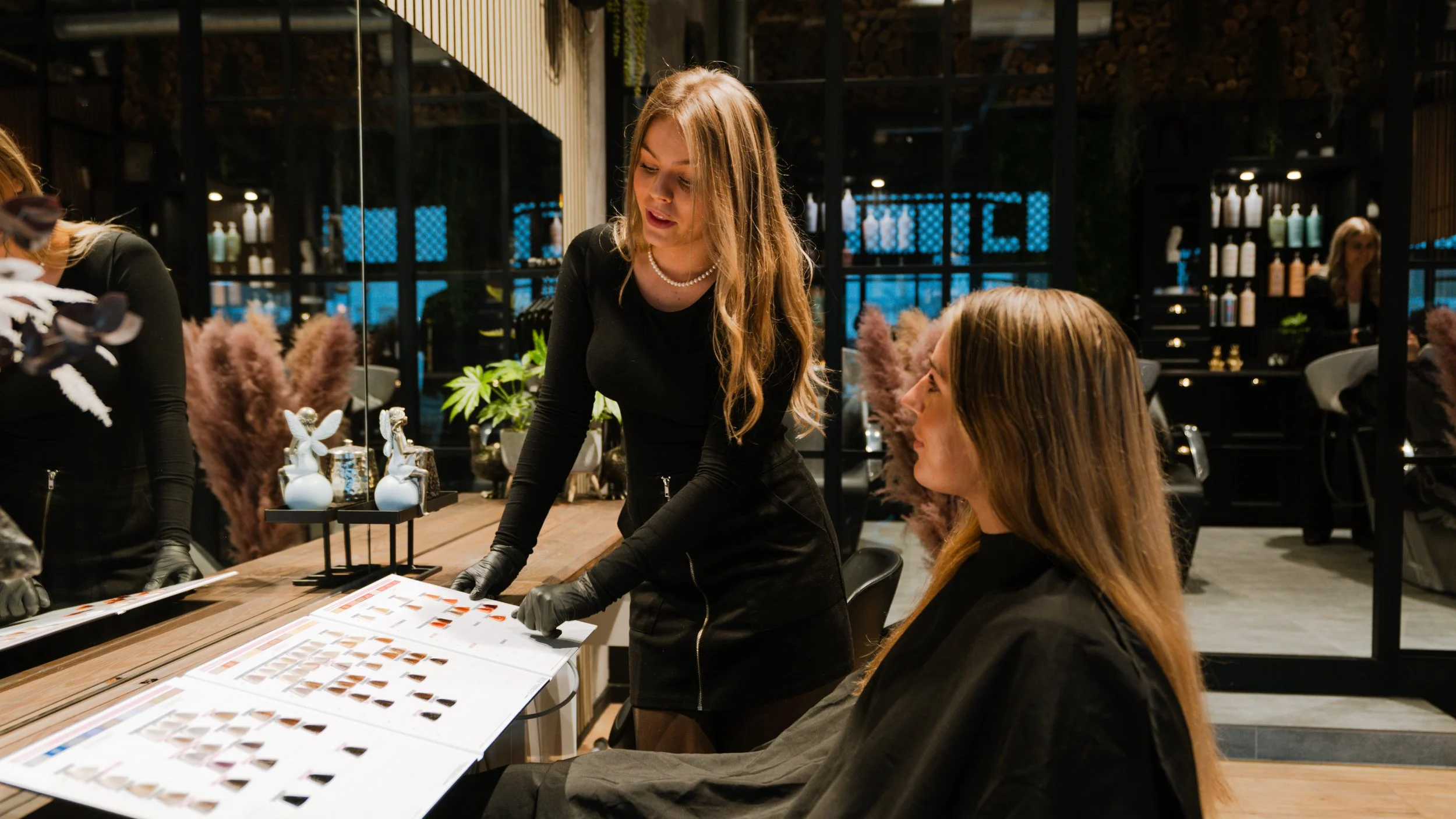 To kvinder snakker i en stilfuld frisørsalon med moderne indretning, spejle, planter og farverige hårprodukter