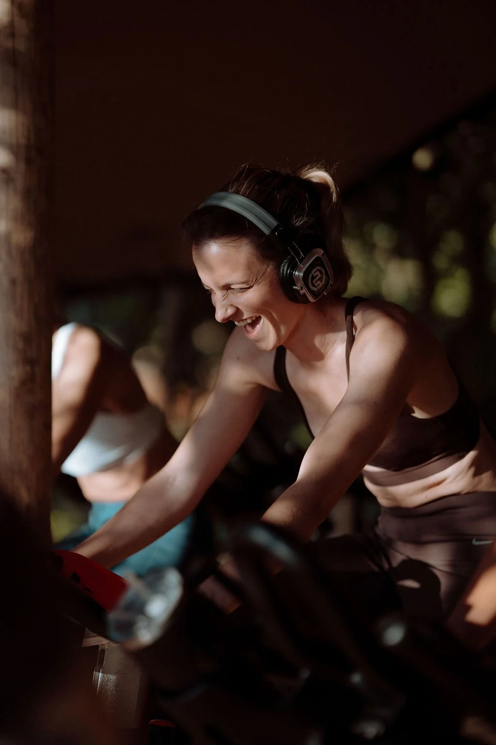 A woman with short dark hair and headphones smiling while exercising on a stationary bike outdoors, with a blurred background of trees and another person exercising.