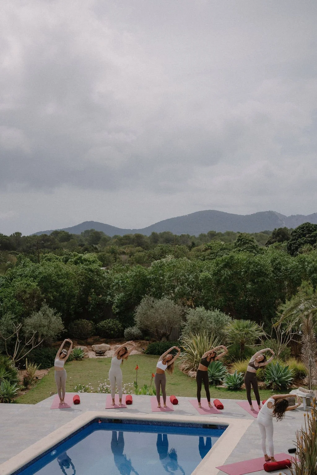 A group of women practicing yoga outdoors beside a swimming pool, with mountains and cloudy sky in the background.