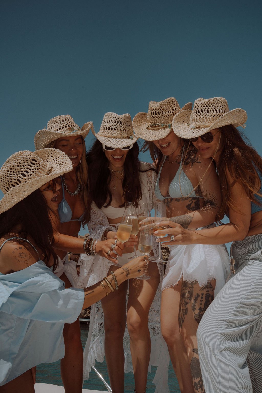 A group of five women in swimwear and lace cover-ups, wearing straw hats and sunglasses, celebrating with champagne on a boat under a clear blue sky.