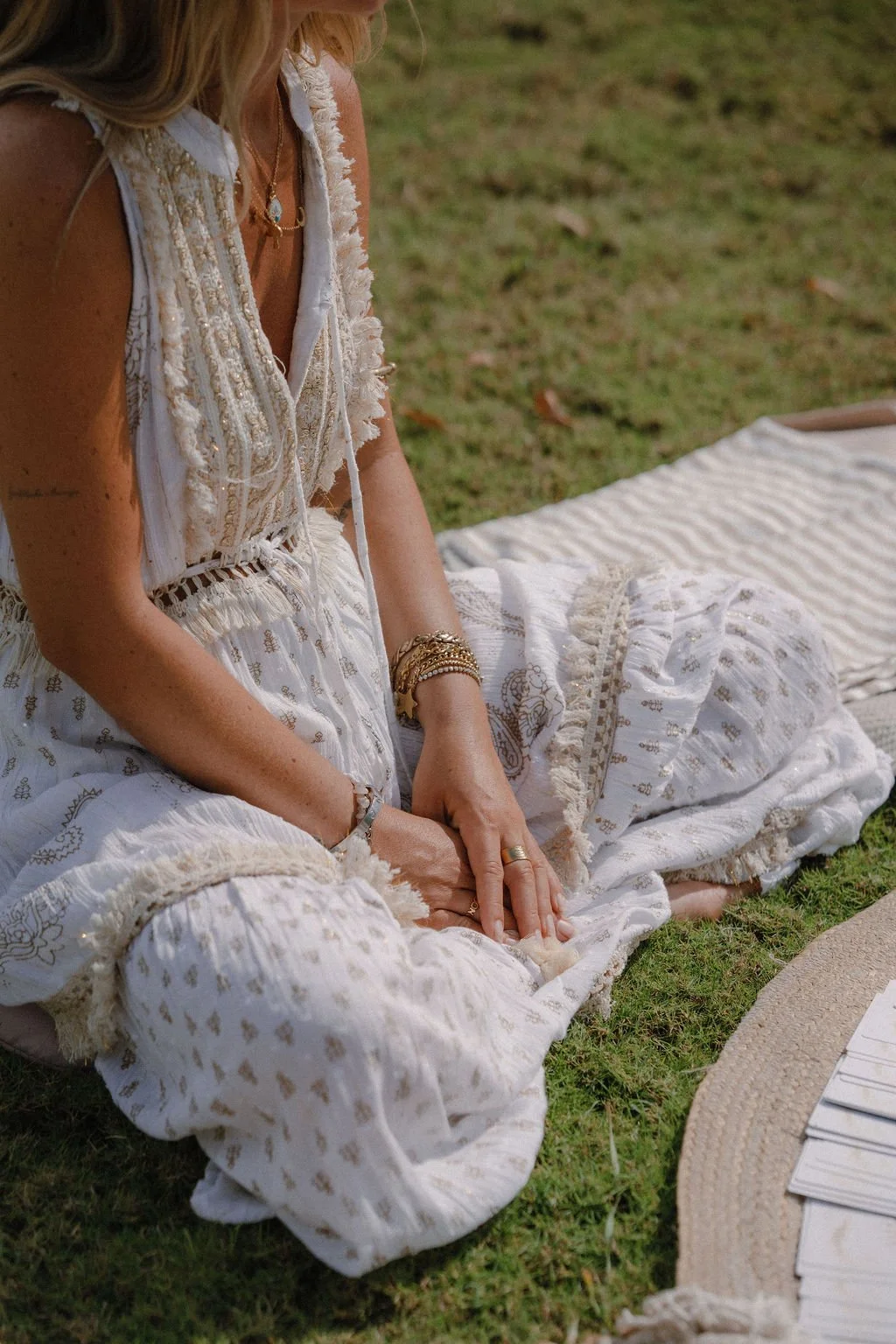 A woman wearing bohemian style white patterned pants and a sleeveless top with embroidery, sitting on grass with her hands resting on her lap, surrounded by a straw hat and other objects.
