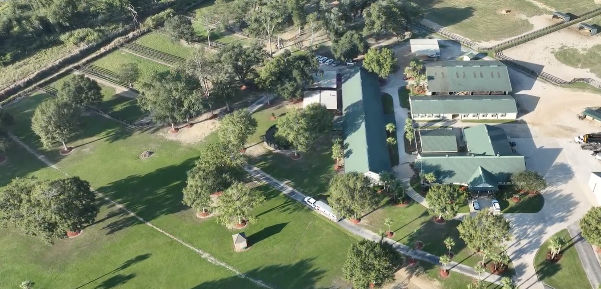 Aerial view of a lush green park with trees, pathways, and a few buildings with green roofs. There are parking areas with cars and a bus, and the park features open grassy areas and small structures.