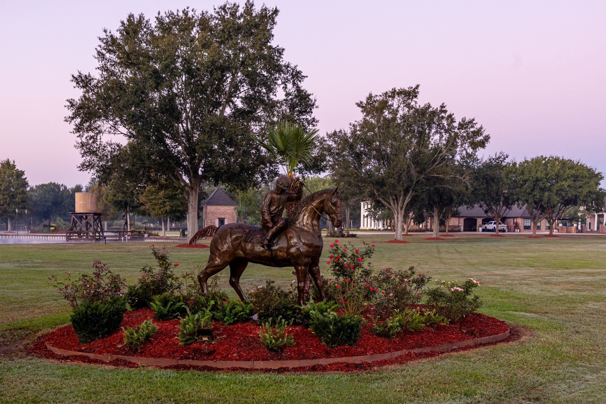Bronze statue of a horse and rider holding a palm leaf, surrounded by flowers, in a park with trees and houses in the background at dusk.