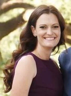 A woman with long, dark hair smiling outdoors, wearing a sleeveless purple dress.