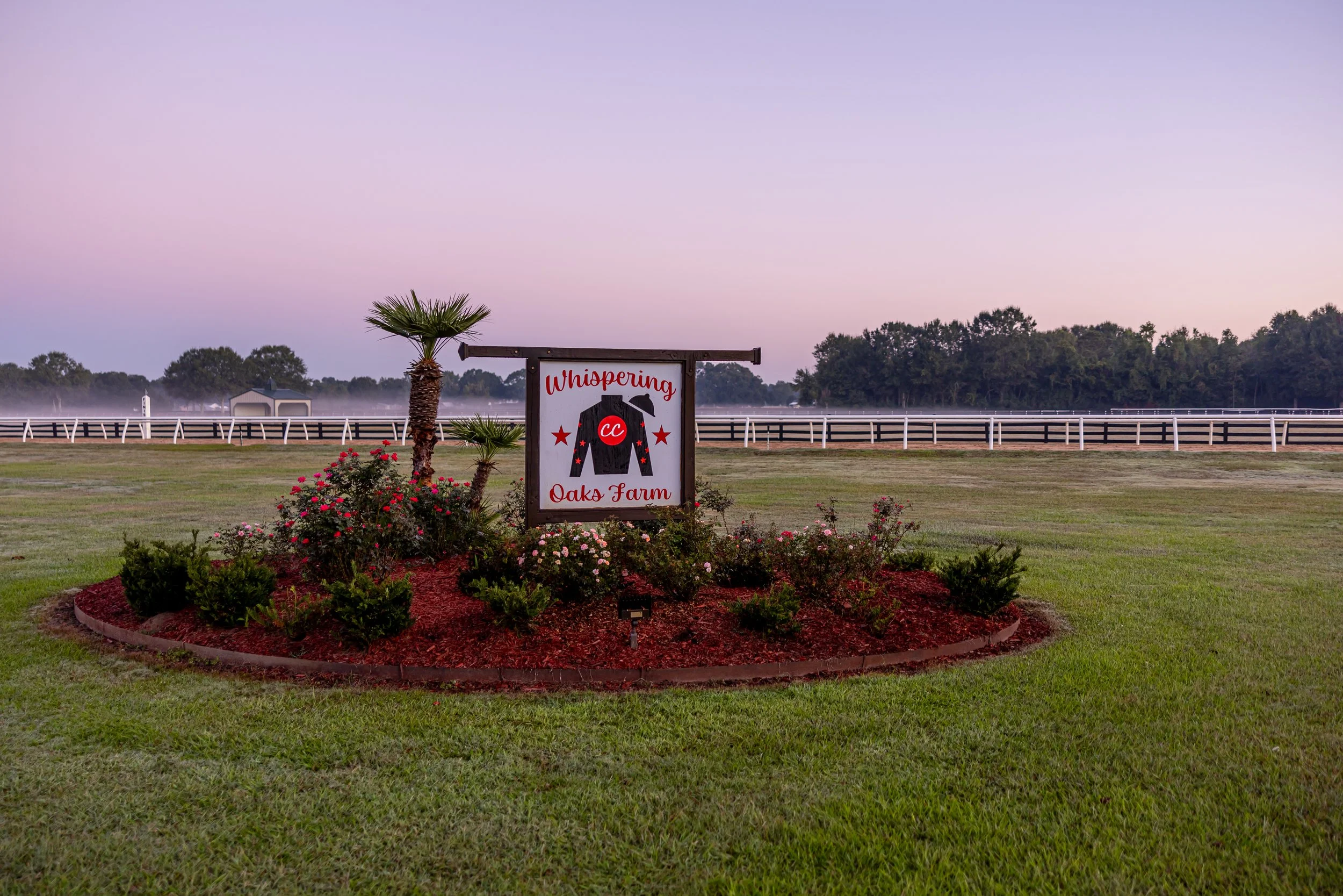 Sign on a farm that reads "Whispering Oako Farm" with a shirt graphic and trees in the background.