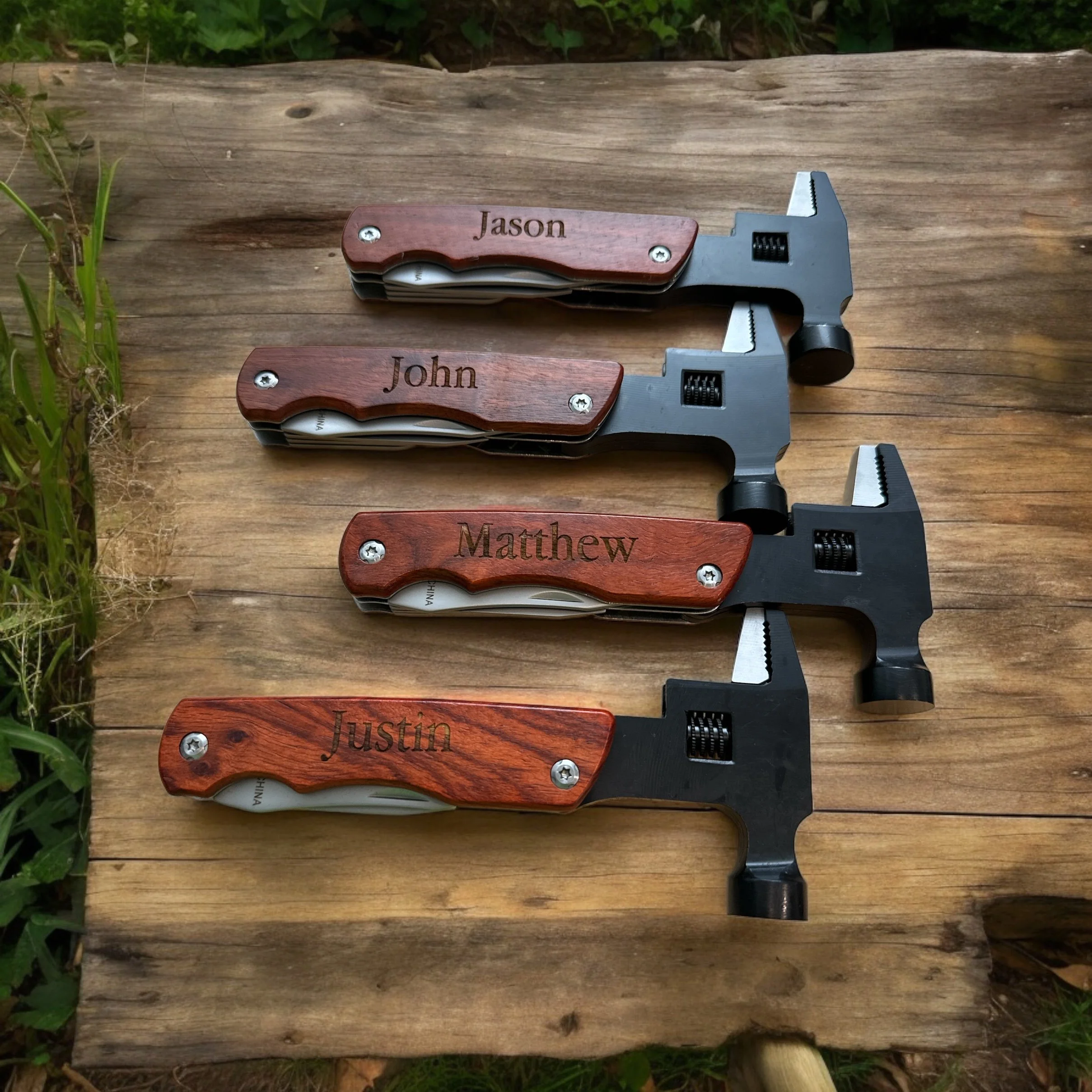 A set of four pocket knives with engraved wooden handles displaying names: Jason, John, Matthew, Justin, on a weathered wooden surface outdoors with green plants nearby.