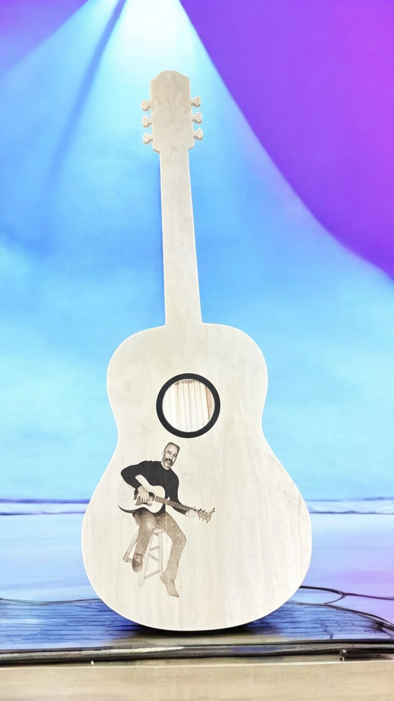 A cardboard cutout shaped like an acoustic guitar with a black and white photo of a man playing guitar at the bottom, a circle cutout near the middle, and a plain headstock at the top, set against a background with colorful stage lighting.