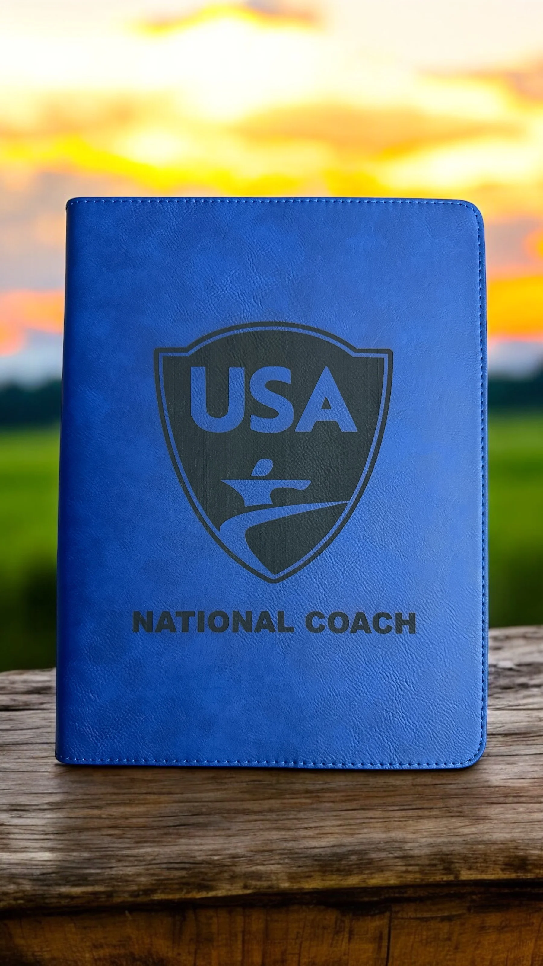 Blue folder with USA national coach emblem, placed on wood surface with outdoor sunset background