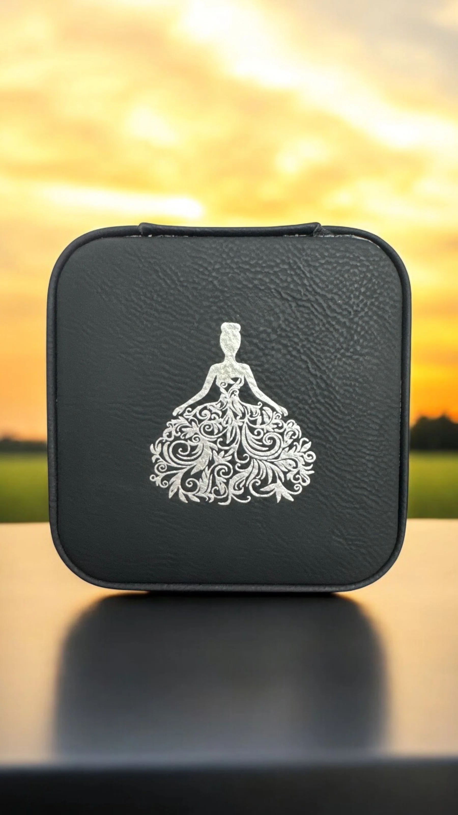 A black box with a silver decorative logo of a woman in a dress with intricate floral patterns, placed outdoors during sunset.
