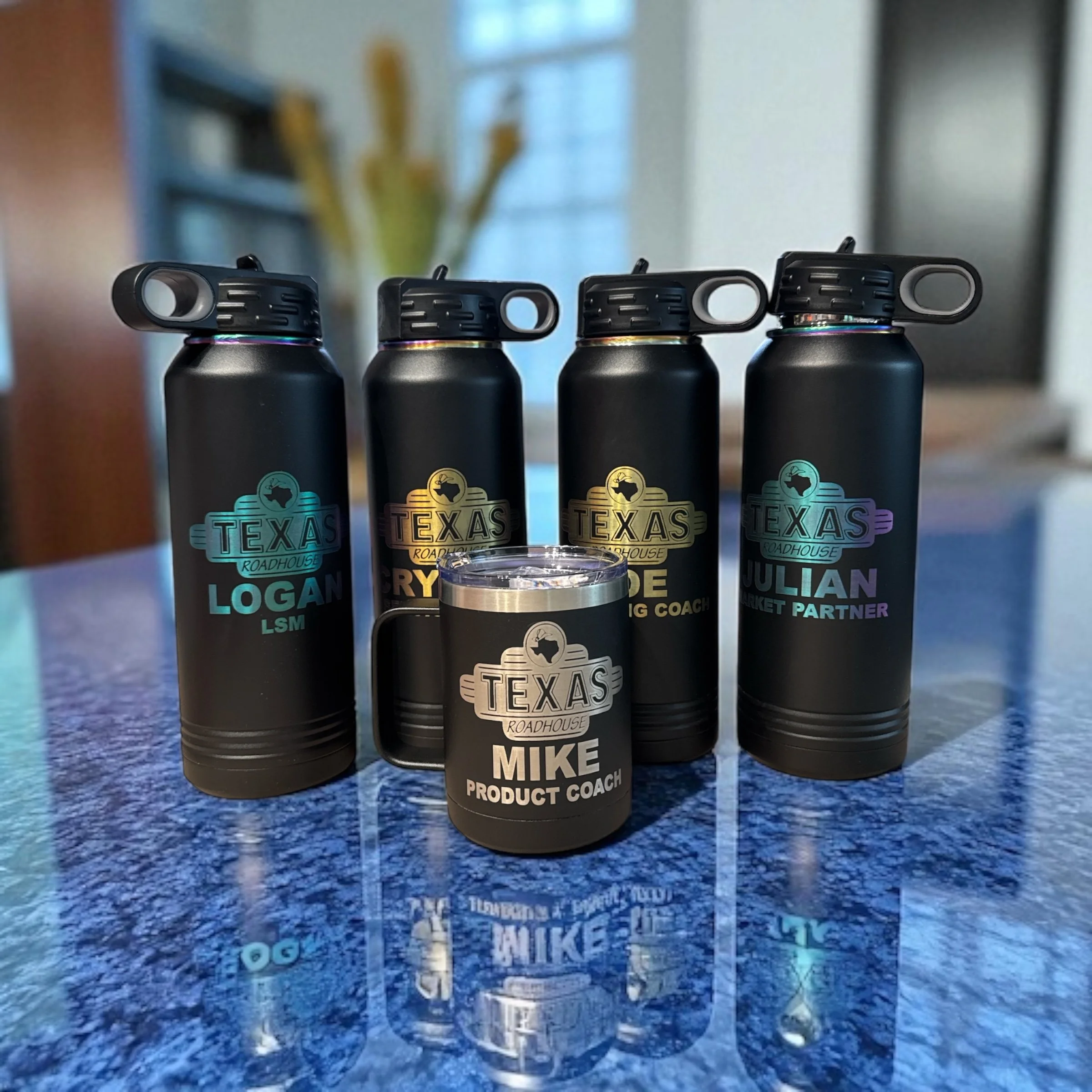 Four black water bottles with colorful labels and a black insulated mug, all placed on a blue speckled countertop. The bottles have various names and titles printed on the labels, including "LOGAN LSM," "MIKE PRODUCT COACH," and others, suggesting th