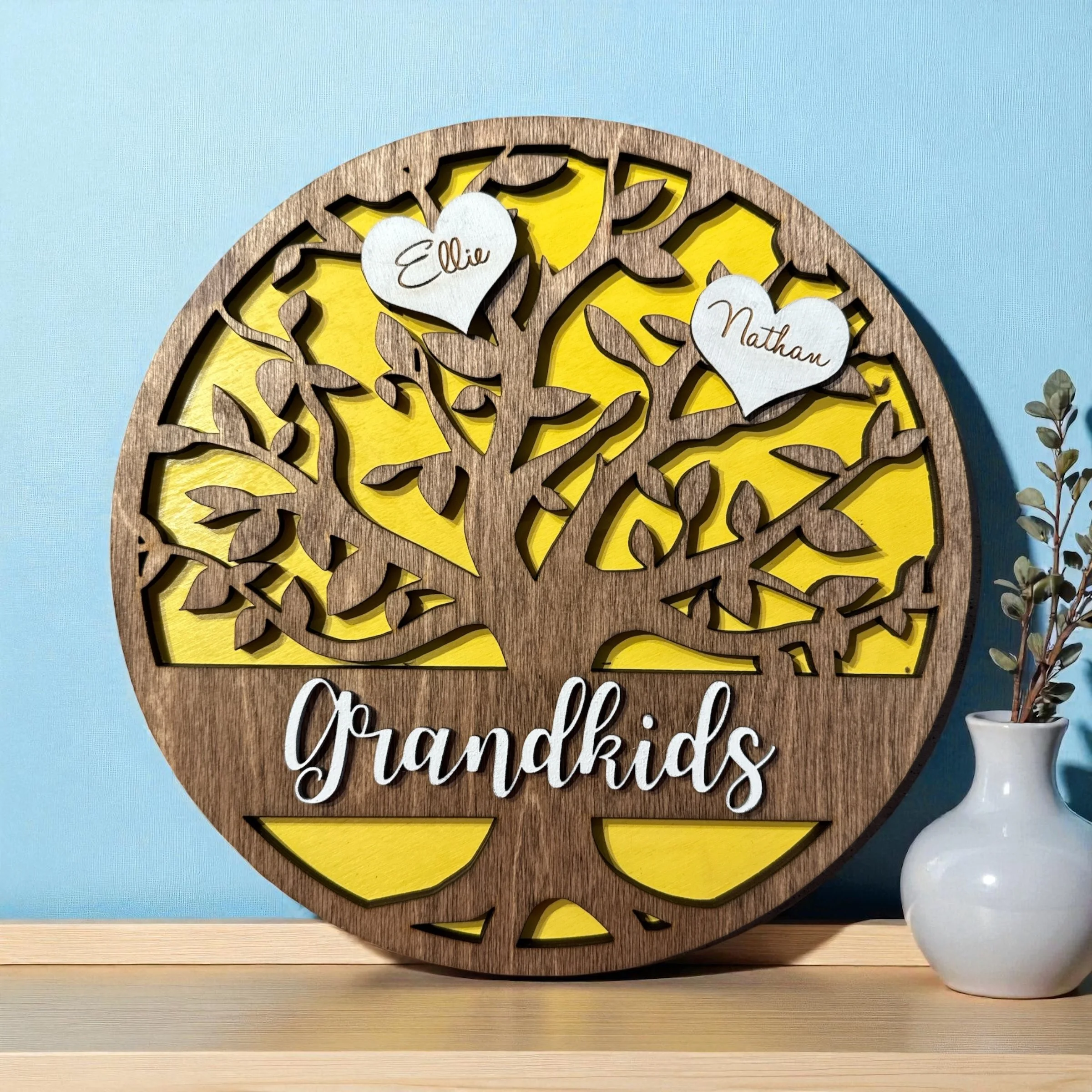 Decorative wall art with a sun motif in wood and yellow, featuring two white heart-shaped signs with the names 'Ellie' and 'Nathan', and the words 'Grateful Gutsy' written at the bottom, placed on a wooden surface next to a white pot with a plant.