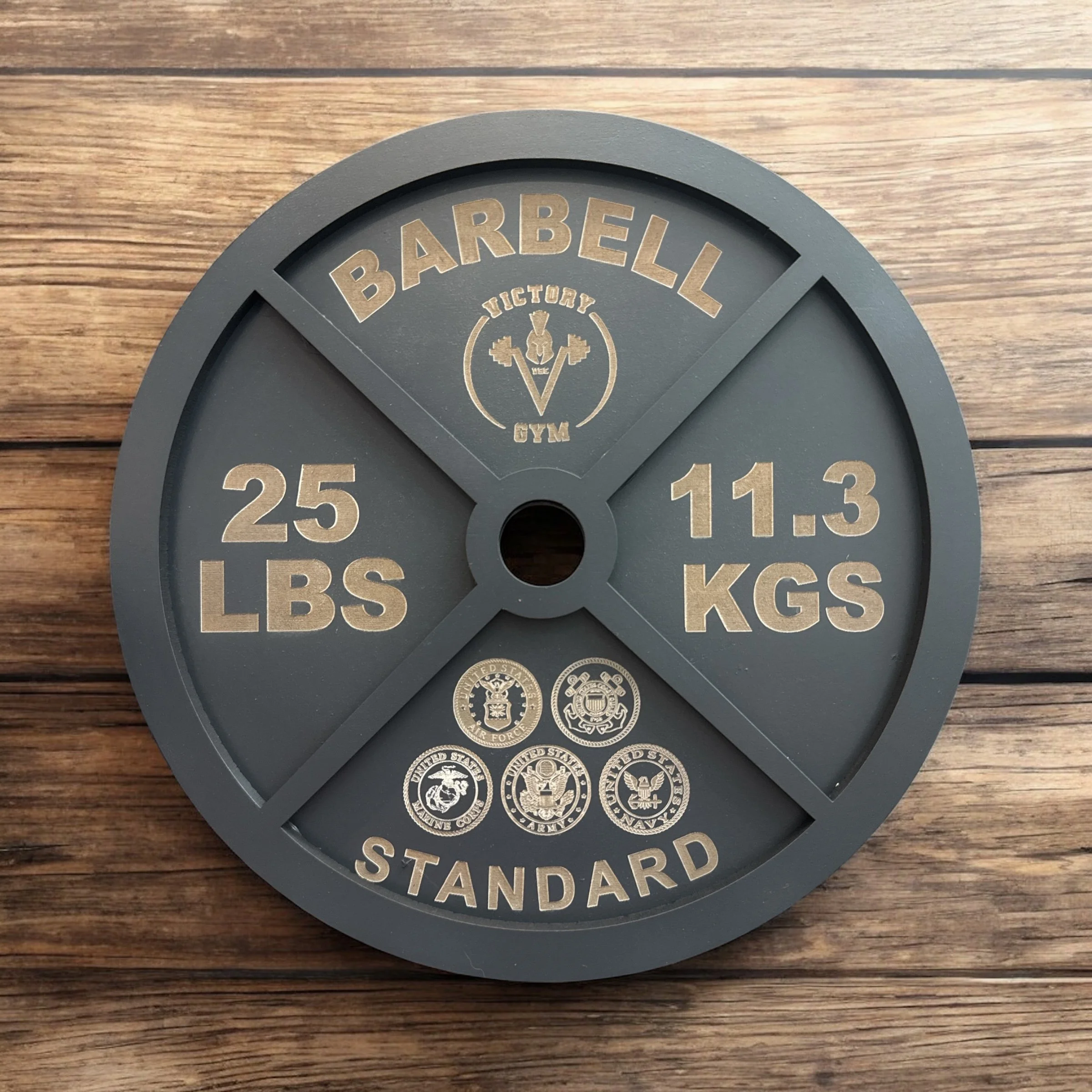 A round, dark gray weight plate from the gym, labeled with the brand "BARBELL GYM" and the word "STANDARD," displaying weights in pounds and kilograms, with official seals at the bottom, placed on a wooden surface.