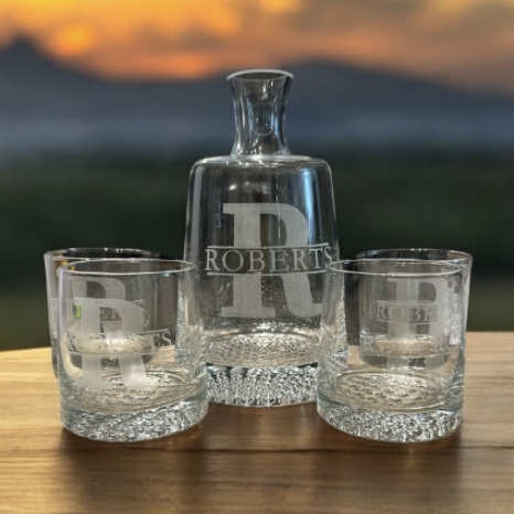 Glass decanter with the initials "R" and the name "Roberts," flanked by two matching whiskey glasses on a wooden surface, with a sunset or sunrise in the background.