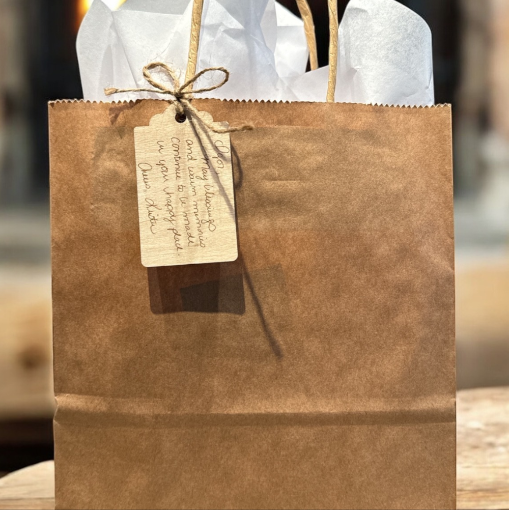 A brown paper gift bag with a gift tag attached by a string bow. Light tissue paper is inside the bag, with handles visible. The background is blurred.