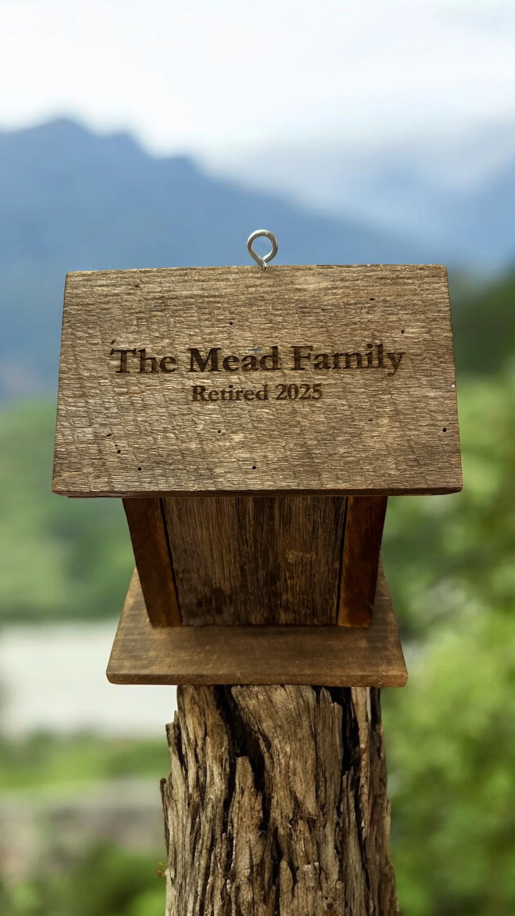 A wooden sign attached to a post, with the text 'The Mead Family Retired 2025', outdoors with blurred background of mountains and greenery.
