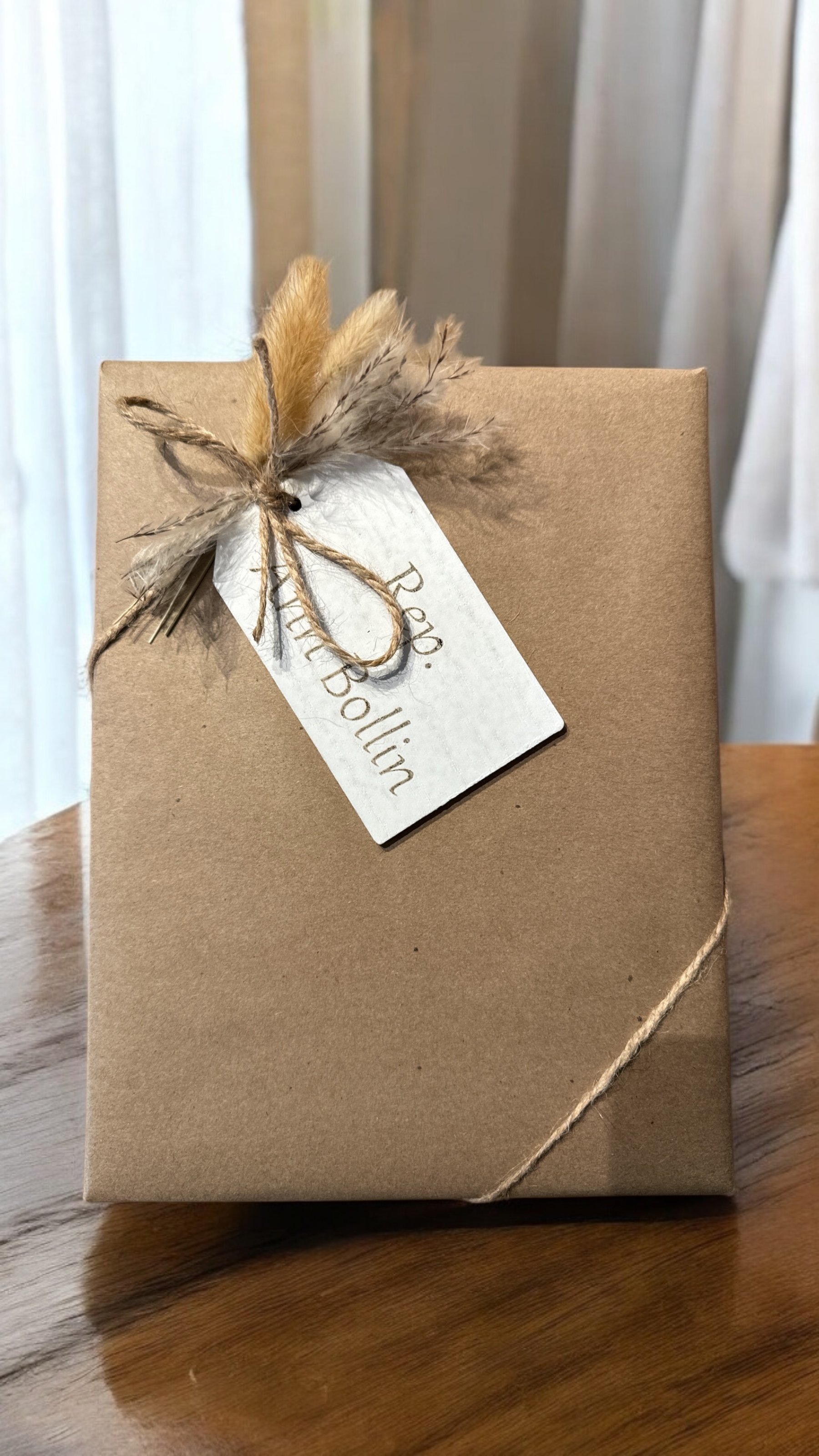 A brown paper gift box decorated with a tag that says 'Reed Bollin', tied with a string, and adorned with a small bunch of dried flowers, placed on a wooden surface with a sheer curtain background.
