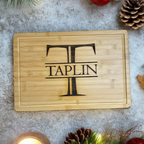 A wooden cutting board with a wine glass and the name 'TAPLIN' engraved on it, surrounded by pine cones and Christmas decorations.