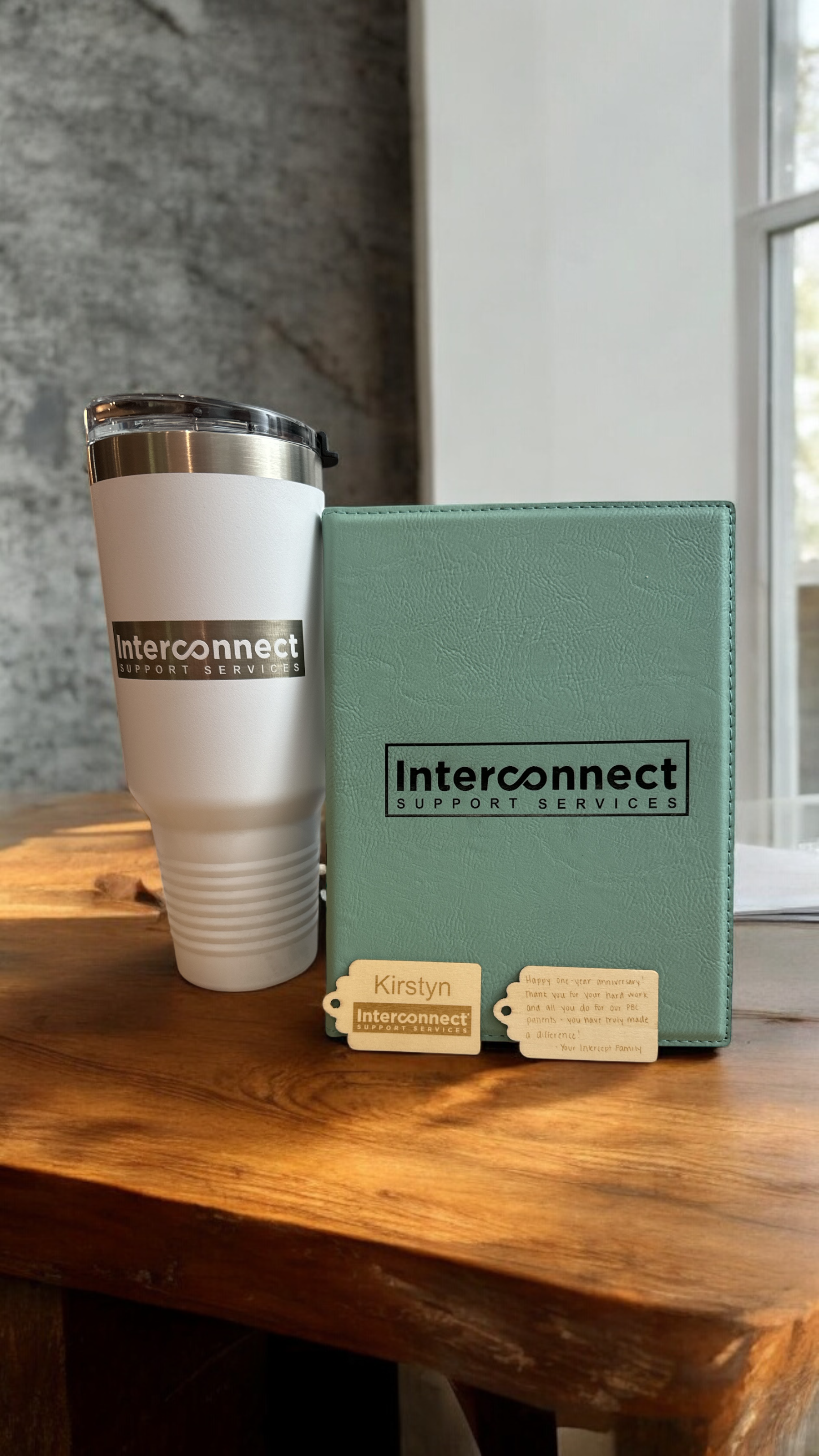 A white tumbler with a black logo and a closed lid, and a light green notebook with black text and wooden tags attached, sitting on a wooden table.