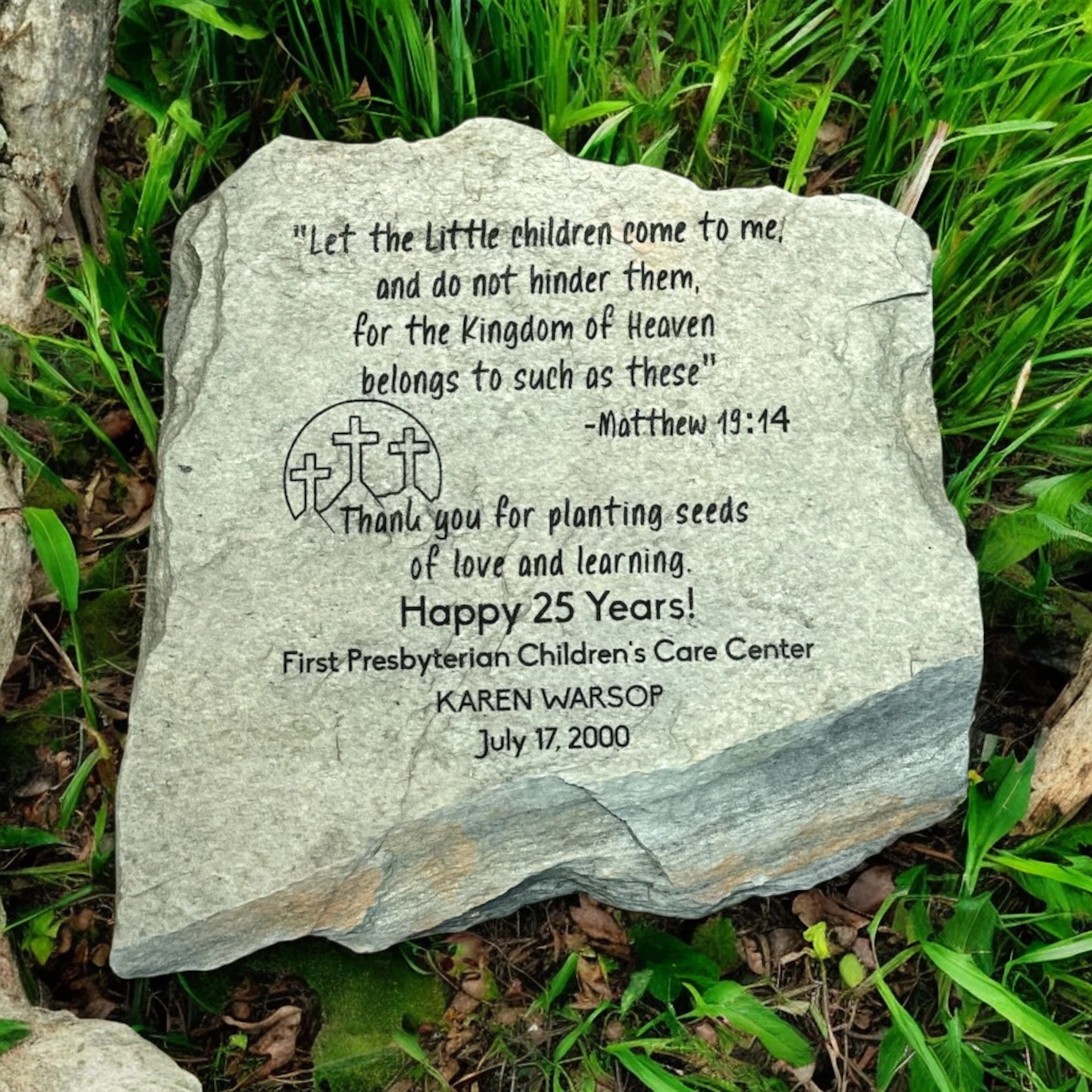 A stone memorial with an engraved quote from Matthew 19:14 resting among green grass and plants.