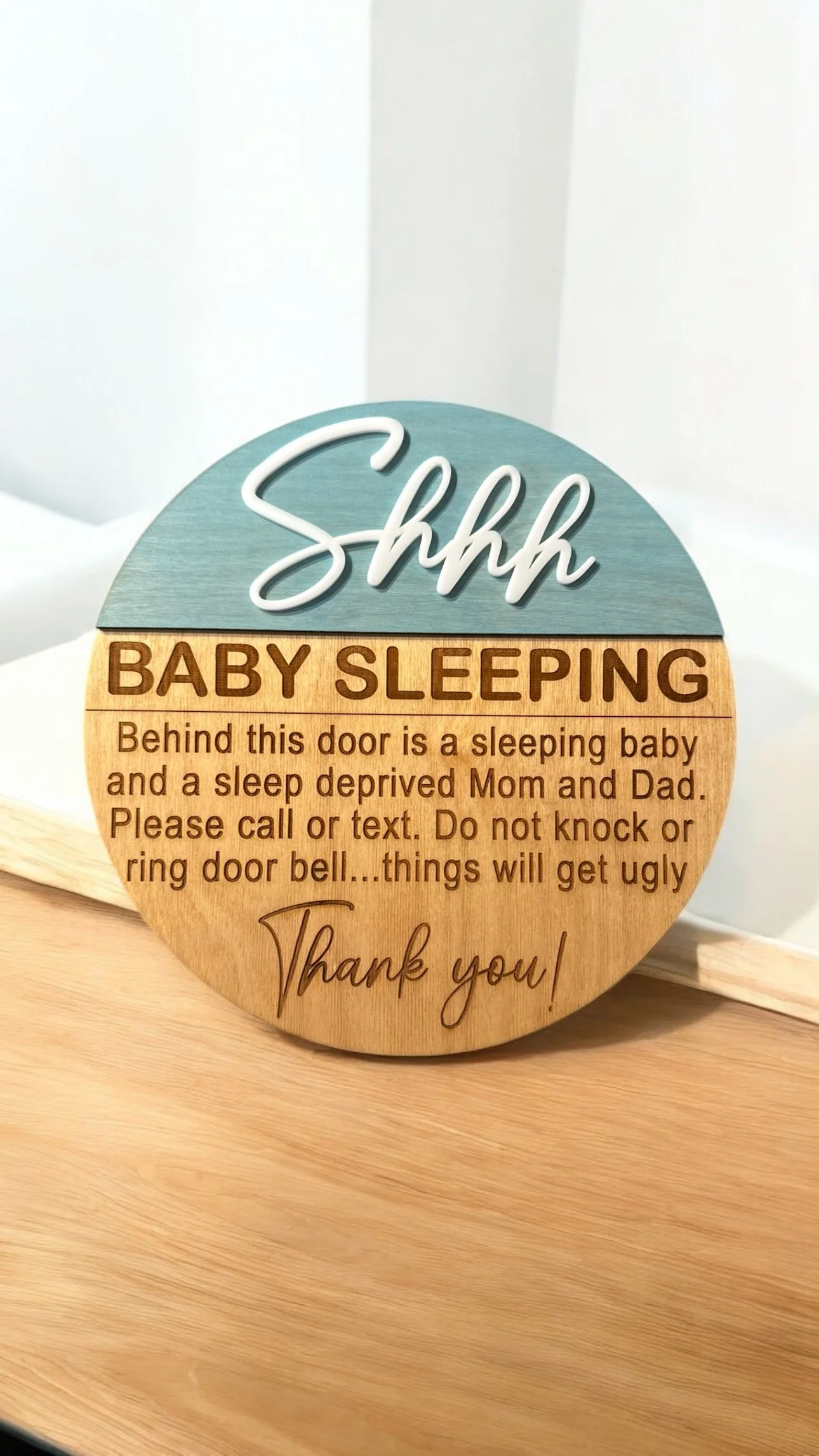 Wooden sign with the text "Shhh BABY SLEEPING" and additional message about not knocking or ringing the doorbell because things will get ugly. The sign is on a wooden surface with a plain white wall in the background.