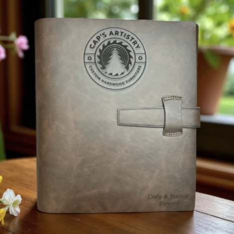 A gray leather-bound journal with a strap closure on a wooden table, with potted plants and flowers in the background.