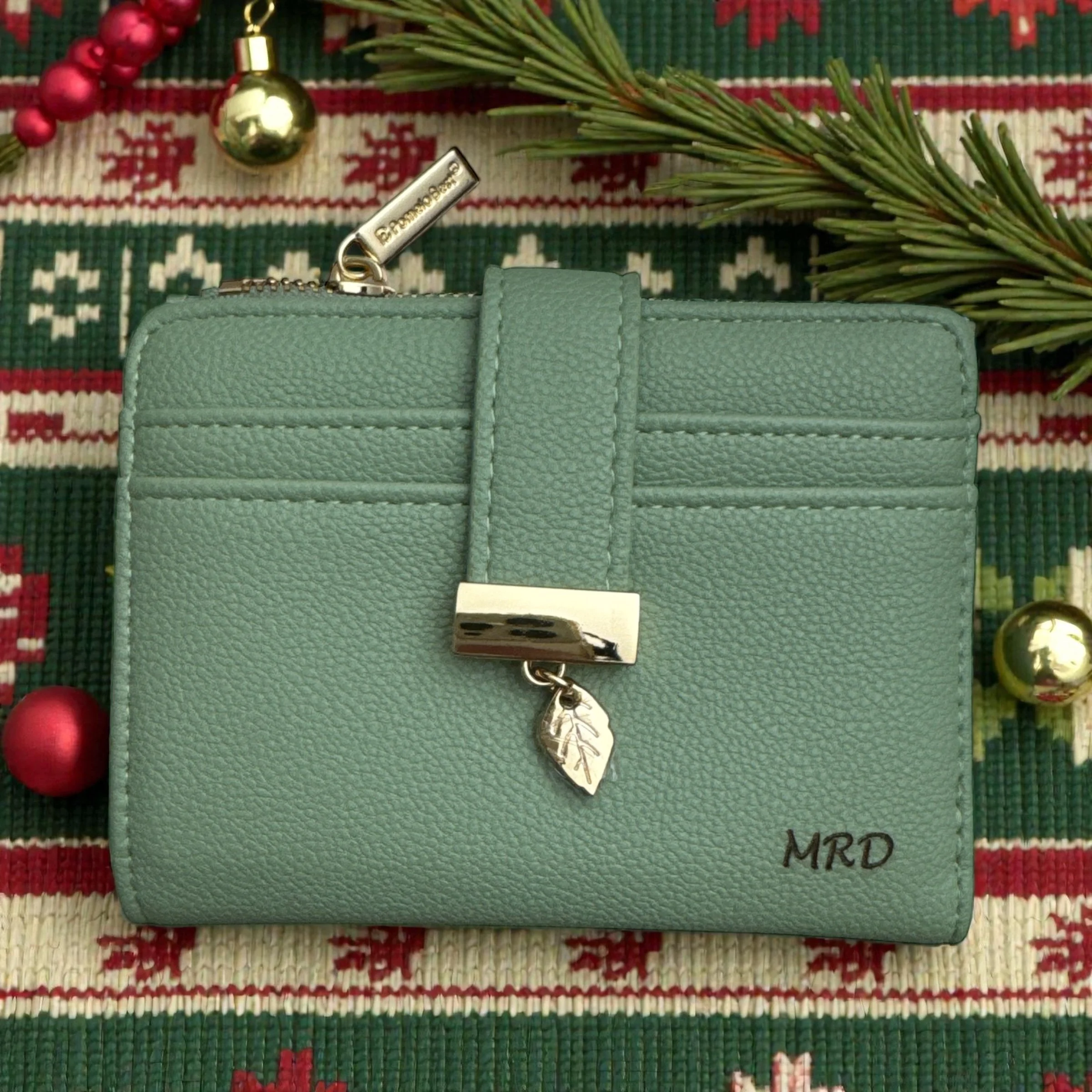 A green wallet with a metal tag attached to a chain, a gold ornament, a red ornament bead, and a sprig of pine leaves on a holiday tablecloth with red, white, and green patterns.