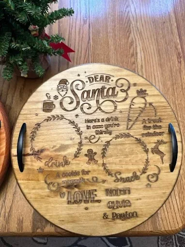 Round wooden serving tray with engraved holiday-themed text and designs, including Santa, a Christmas tree, a wrapped gift, and a reindeer, placed on a wooden floor with part of a Christmas tree visible in the corner.