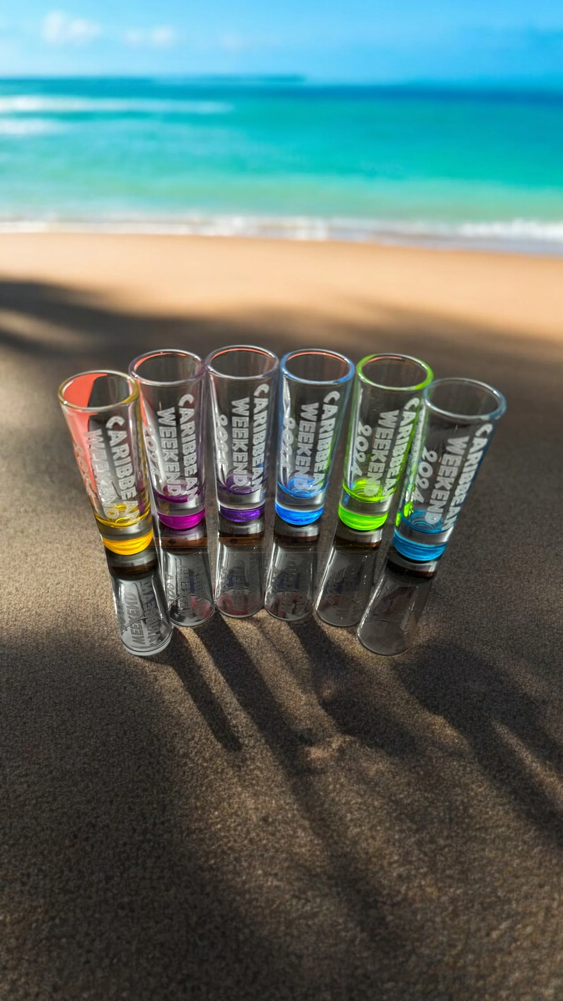 Six colorful shot glasses on a sandy beach with a turquoise ocean and blue sky in the background.