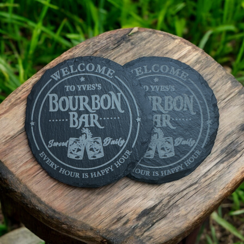 Two round black coasters on a wooden surface, each with a vintage-style bourbon bar design and text welcoming guests, with grass in the background.