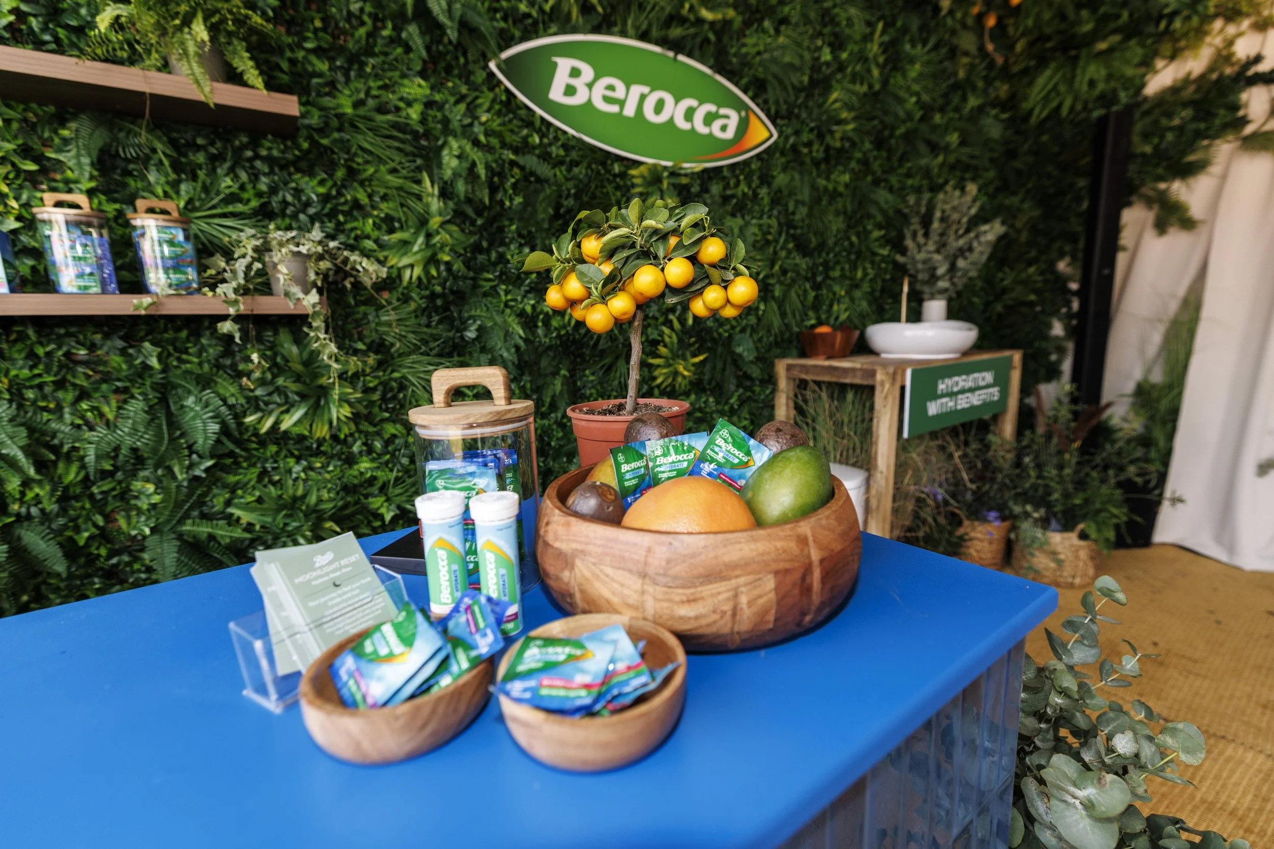 A display of Berocca energy vitamins and health products on a blue table, with a small potted orange tree, and a lush green vertical garden wall with a Berocca sign in the background.