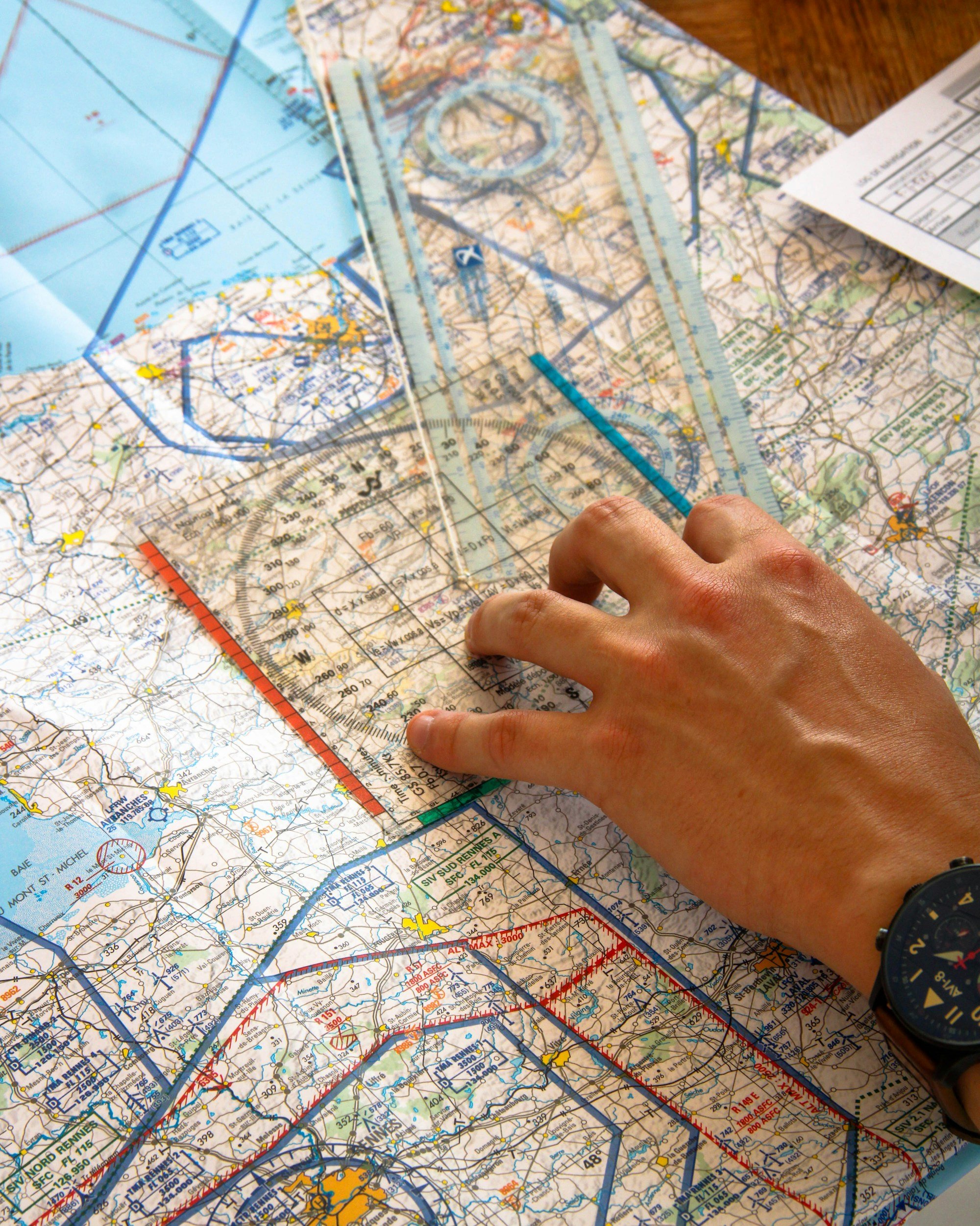Person's hand measuring a detailed road map with a clear plastic ruler, with multiple maps and papers laid out on a wooden table.