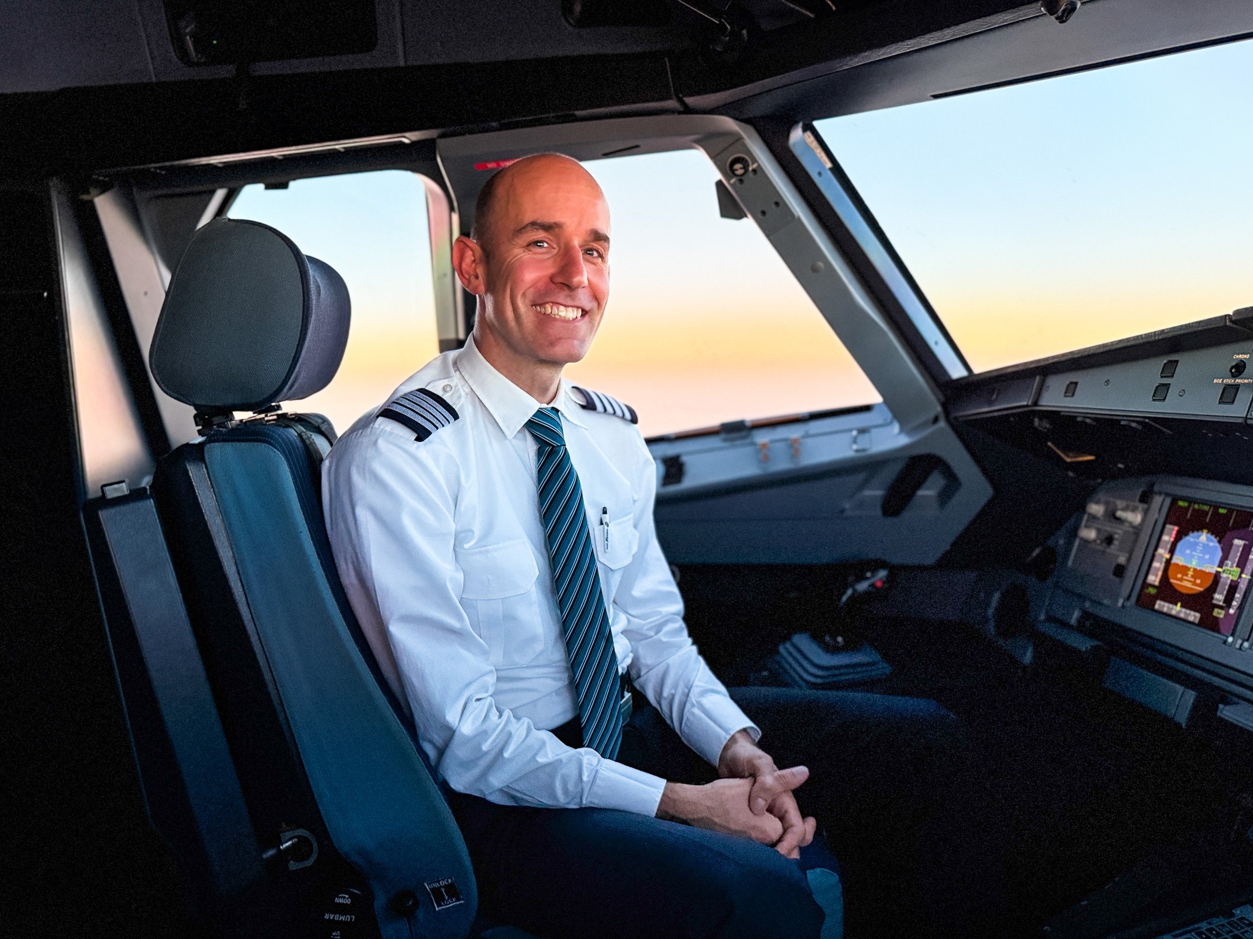 A smiling pilot in a white shirt and striped tie sitting in the cockpit of an airplane at sunset.