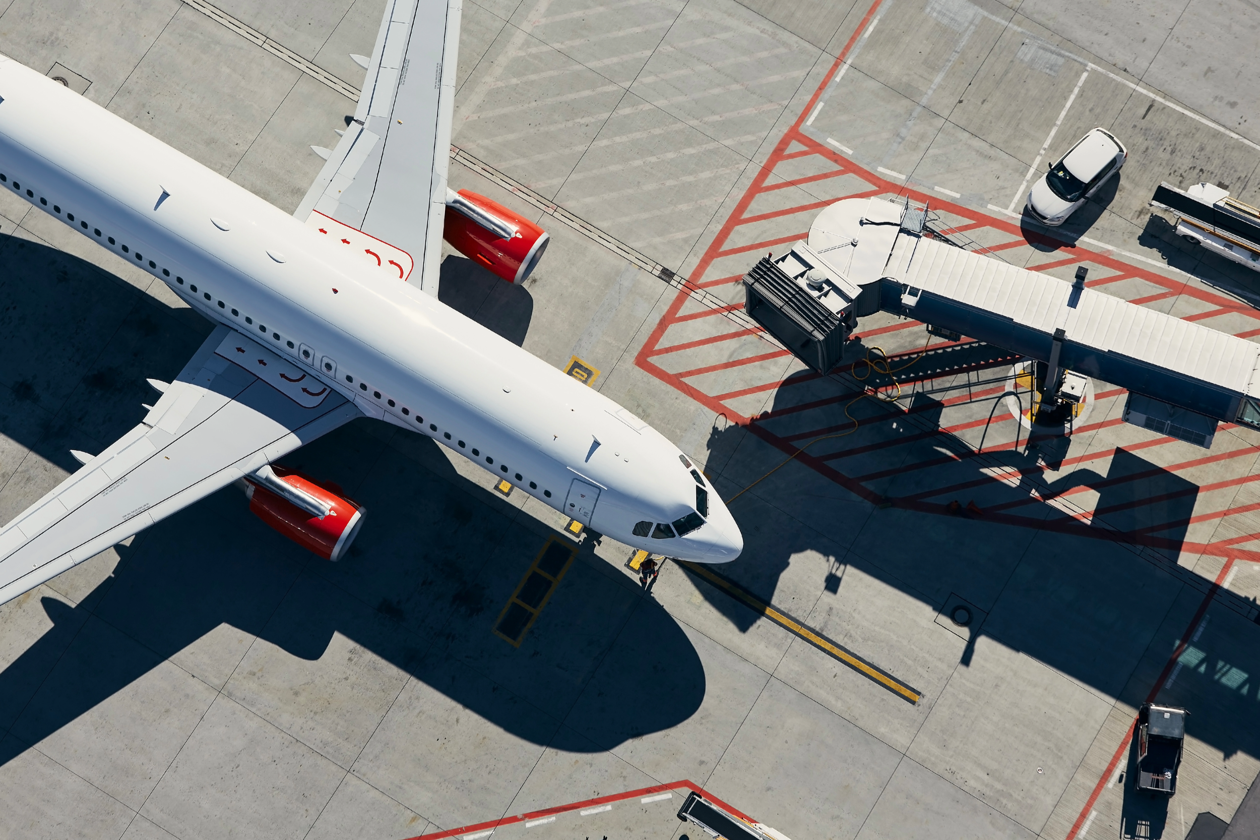Overhead view of a white commercial airplane parked at a gate, connected to a jet bridge, with various vehicles around including a luggage cart and a small car.