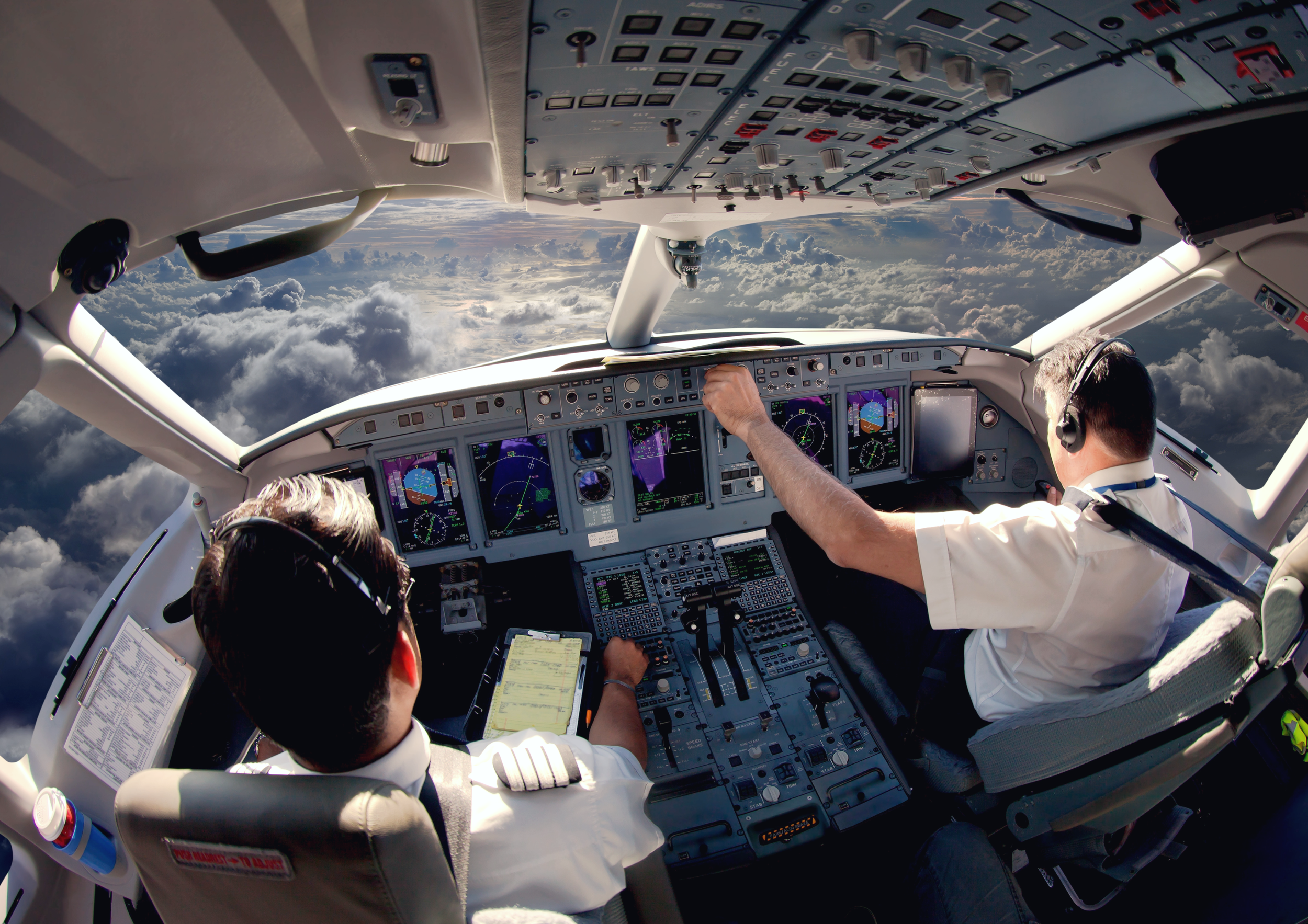 Cockpit view of an airplane with two pilots flying above the clouds, one pilot is adjusting controls while the other is looking at a checklist.
