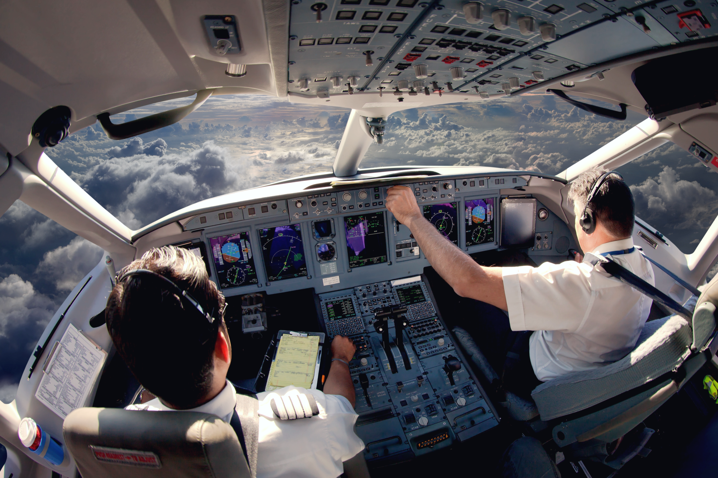 Photo of two pilots in the cockpit of a commercial airplane flying above clouds, with various instruments and controls visible on the dashboard.