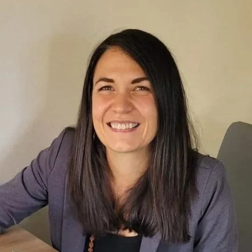 A woman with long dark hair, smiling, wearing a gray blazer, sitting at a table against a plain wall.