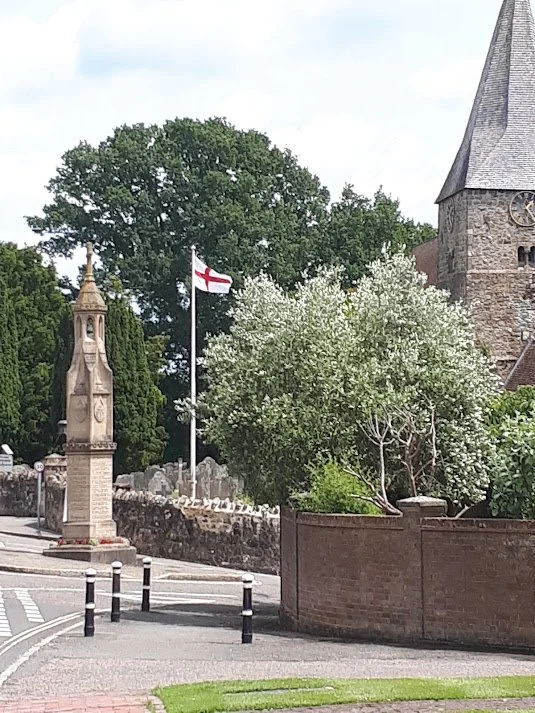 A church with a tall steeple, a flagpole with the English flag, a monument with a cross, and a garden with blooming trees and bushes.